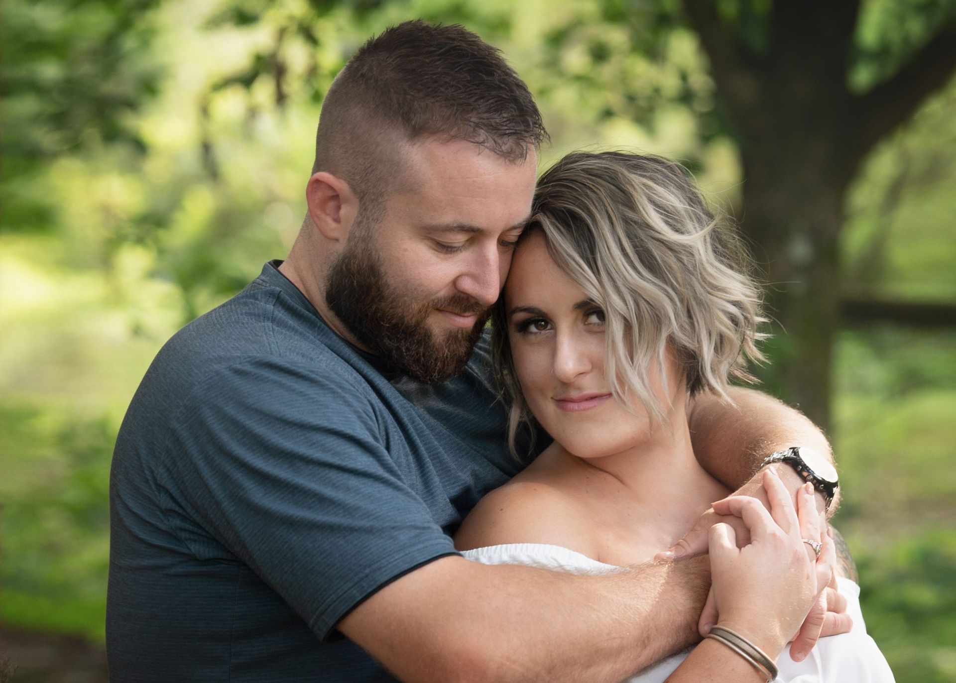 Couple embracing outdoors; man with beard, woman with off-shoulder top; soft focus greenery in background.