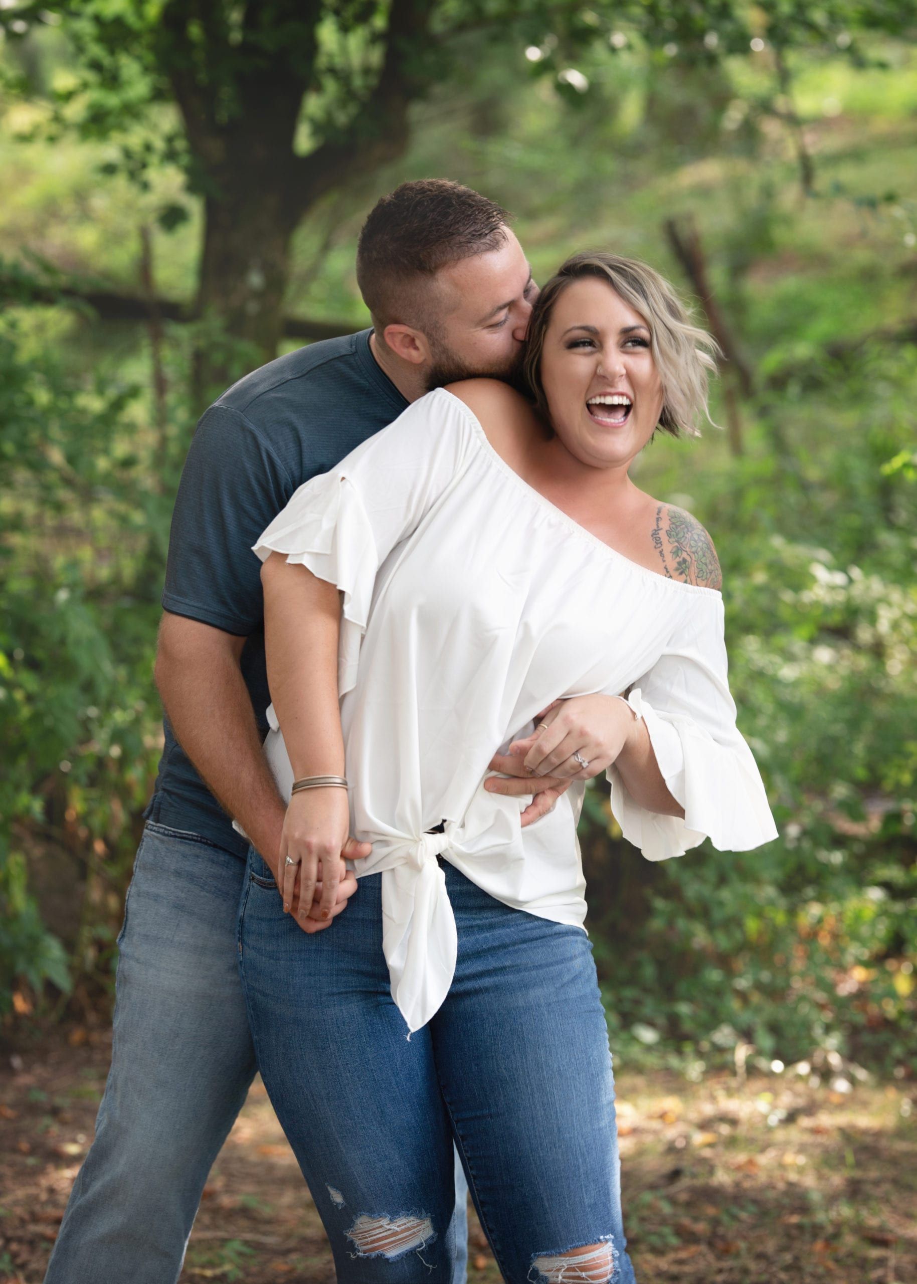Man kissing woman's cheek, both laughing in a wooded area.