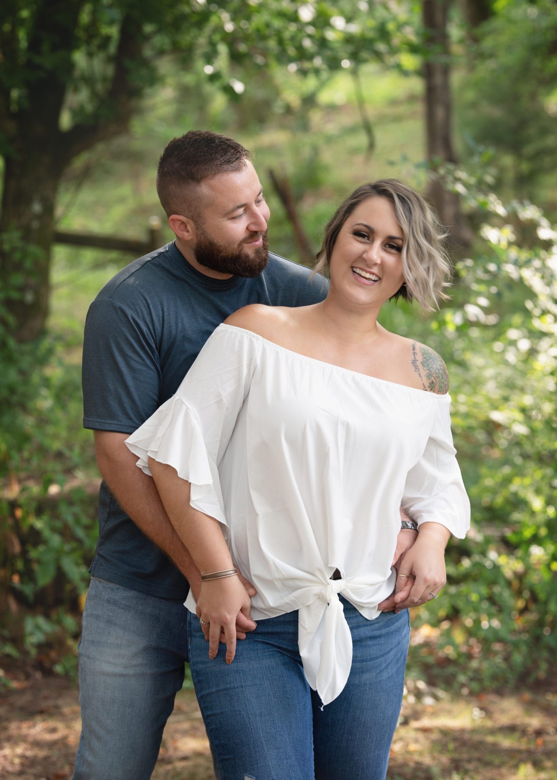 Man and woman smiling, embracing outdoors. He looks at her; she's in a white off-the-shoulder top, jeans. Trees in background.