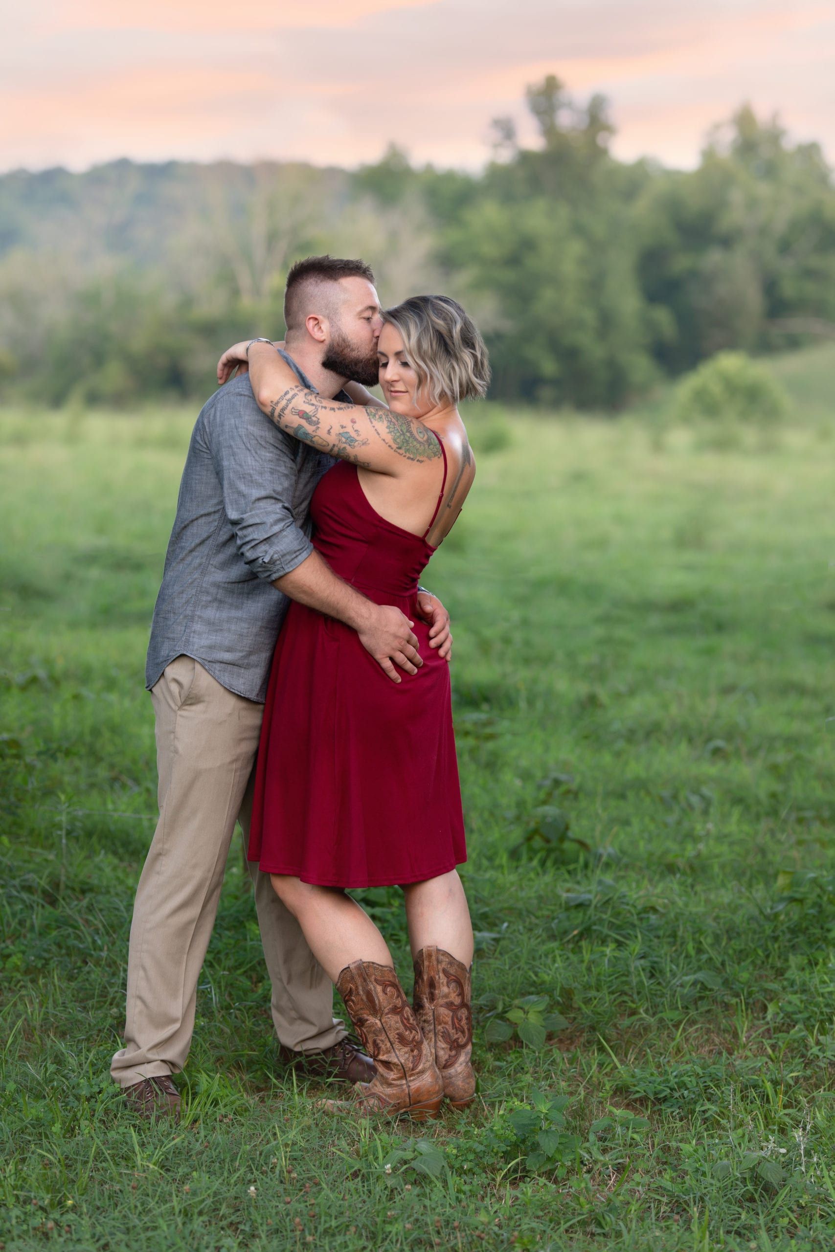 Couple embraces in field; man kisses woman's forehead. She wears red dress, cowboy boots; he has tattoos, button-down shirt.