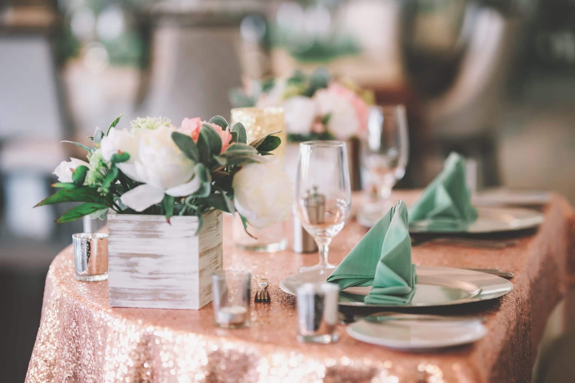 Wedding reception table with floral centerpiece, rose gold tablecloth, and green napkins.