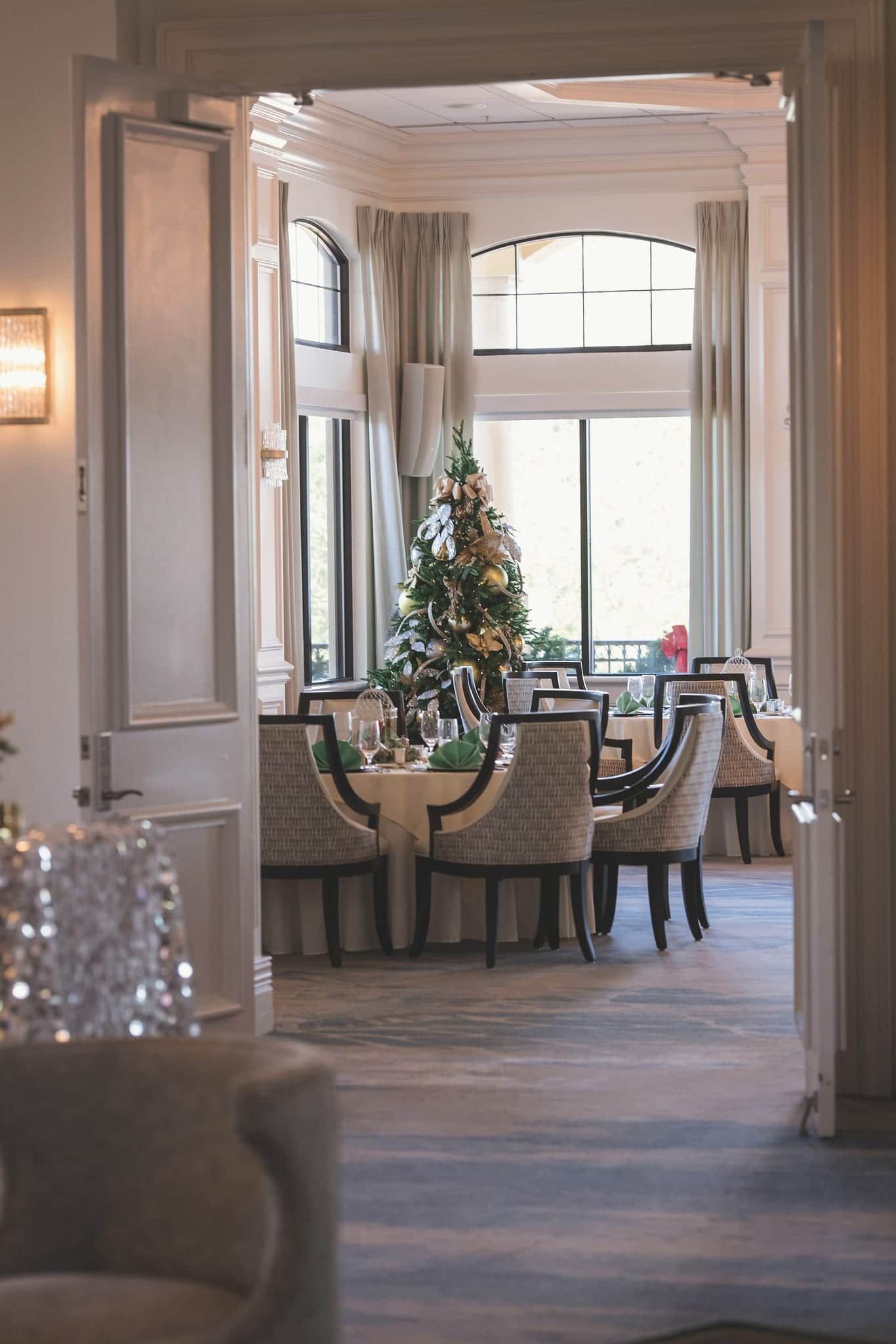 Elegant dining room with Christmas tree, tables set for a meal. Neutral tones with sunlight.