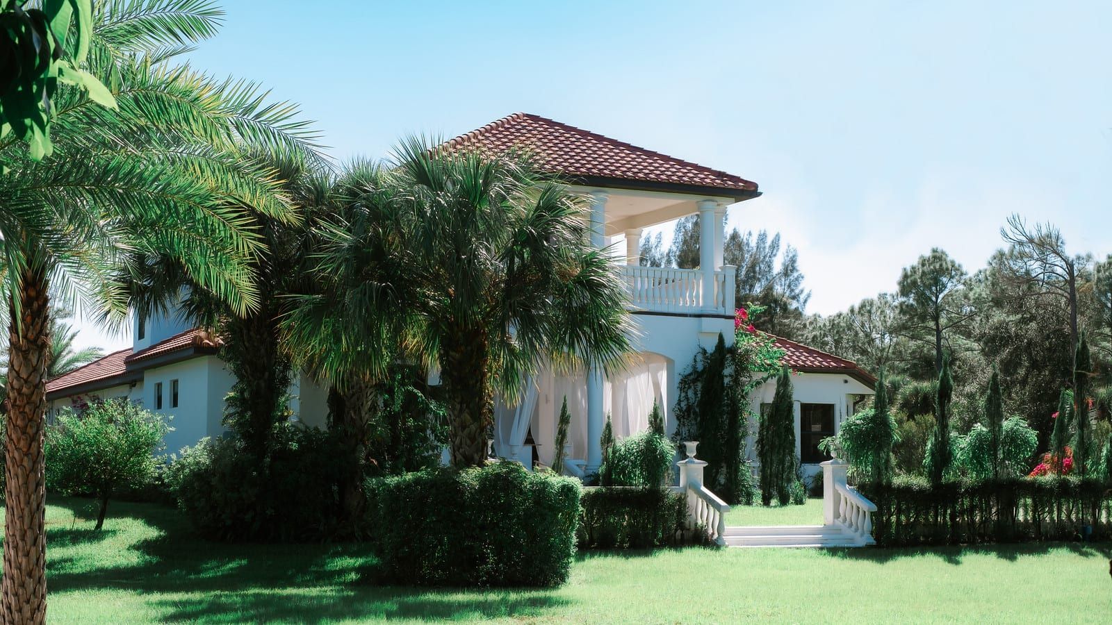 White stucco house with red tile roof, surrounded by green lawn, palm trees, and bushes.