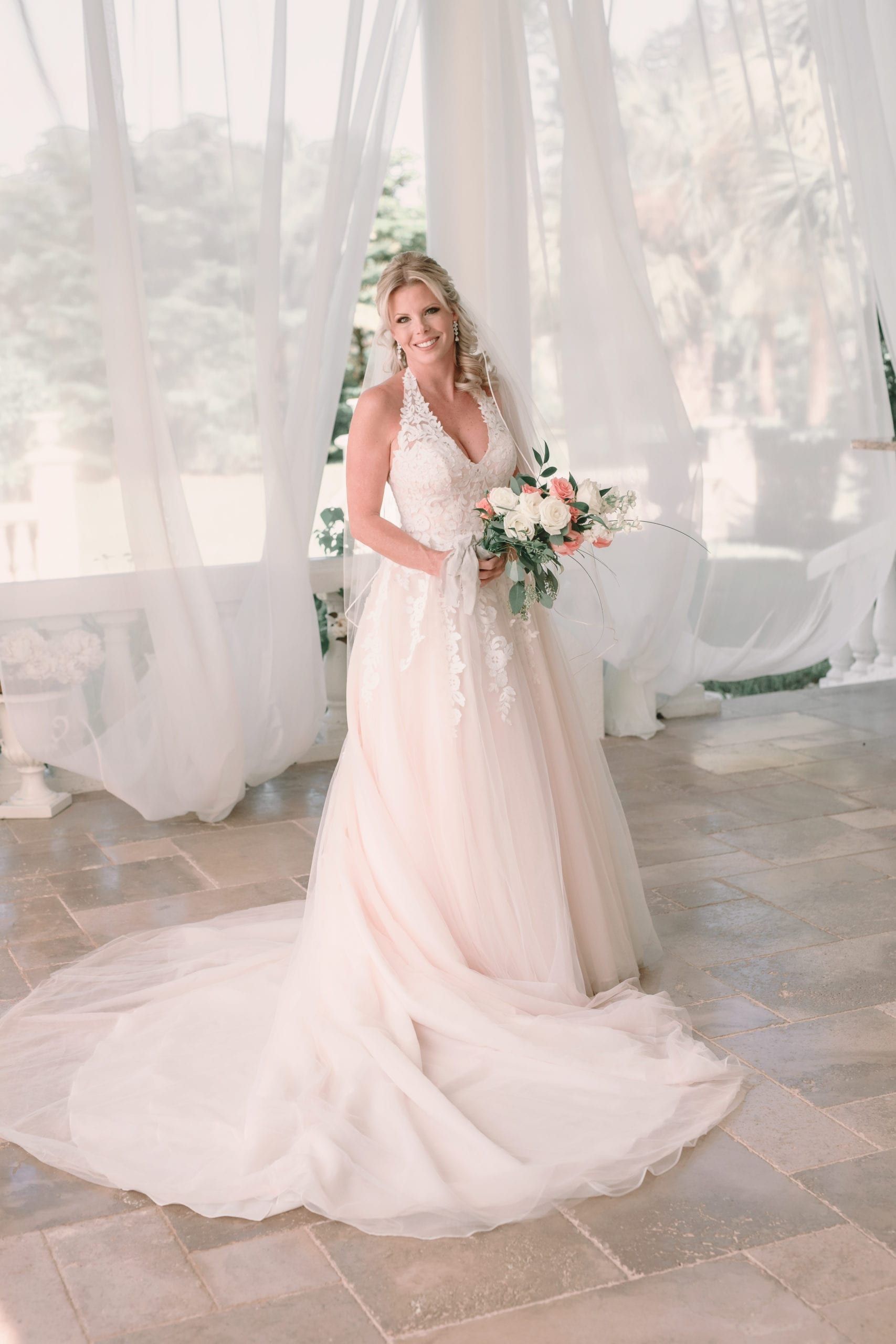 Blonde bride in a white and blush gown holds a bouquet.  She's smiling in a bright, open-air space.