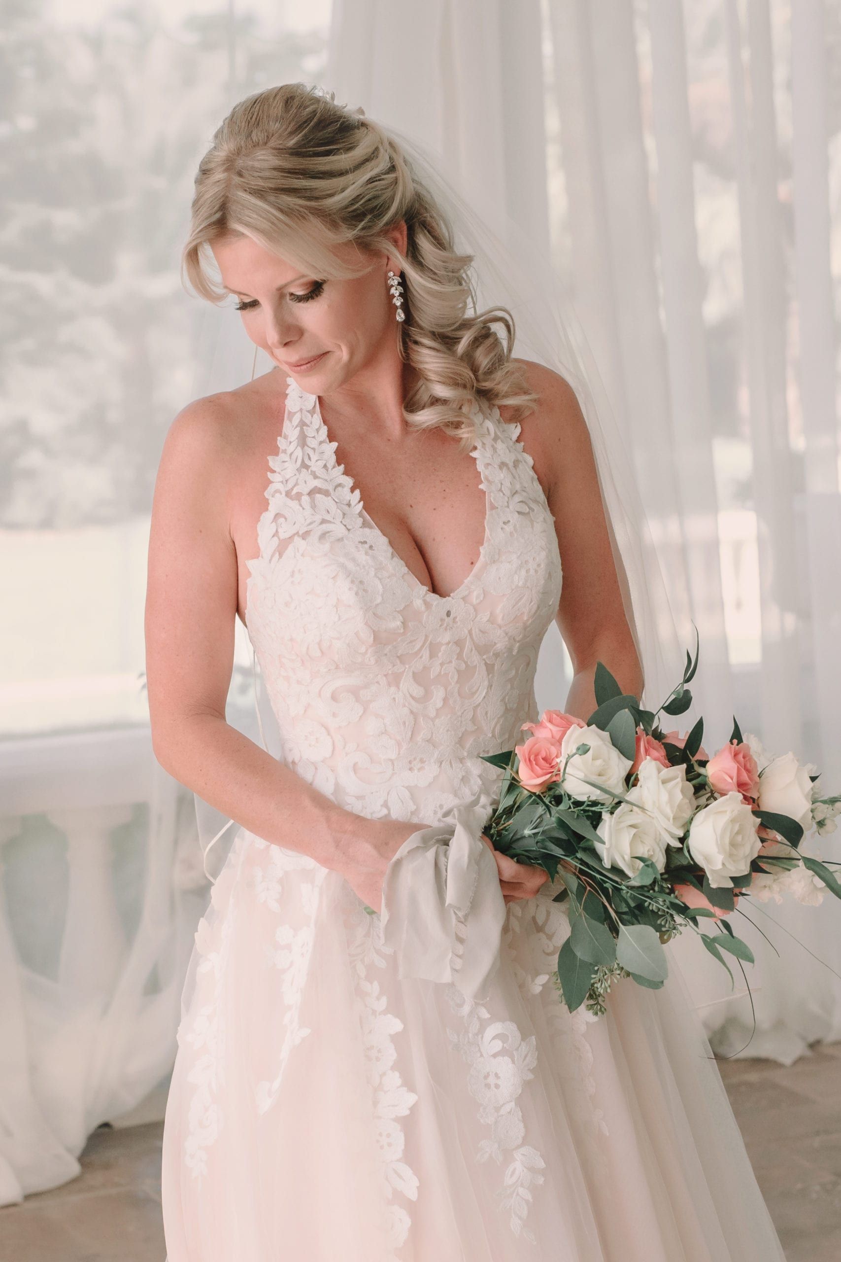 Blonde bride in lace halter dress, holding bouquet, looking down. Soft lighting.
