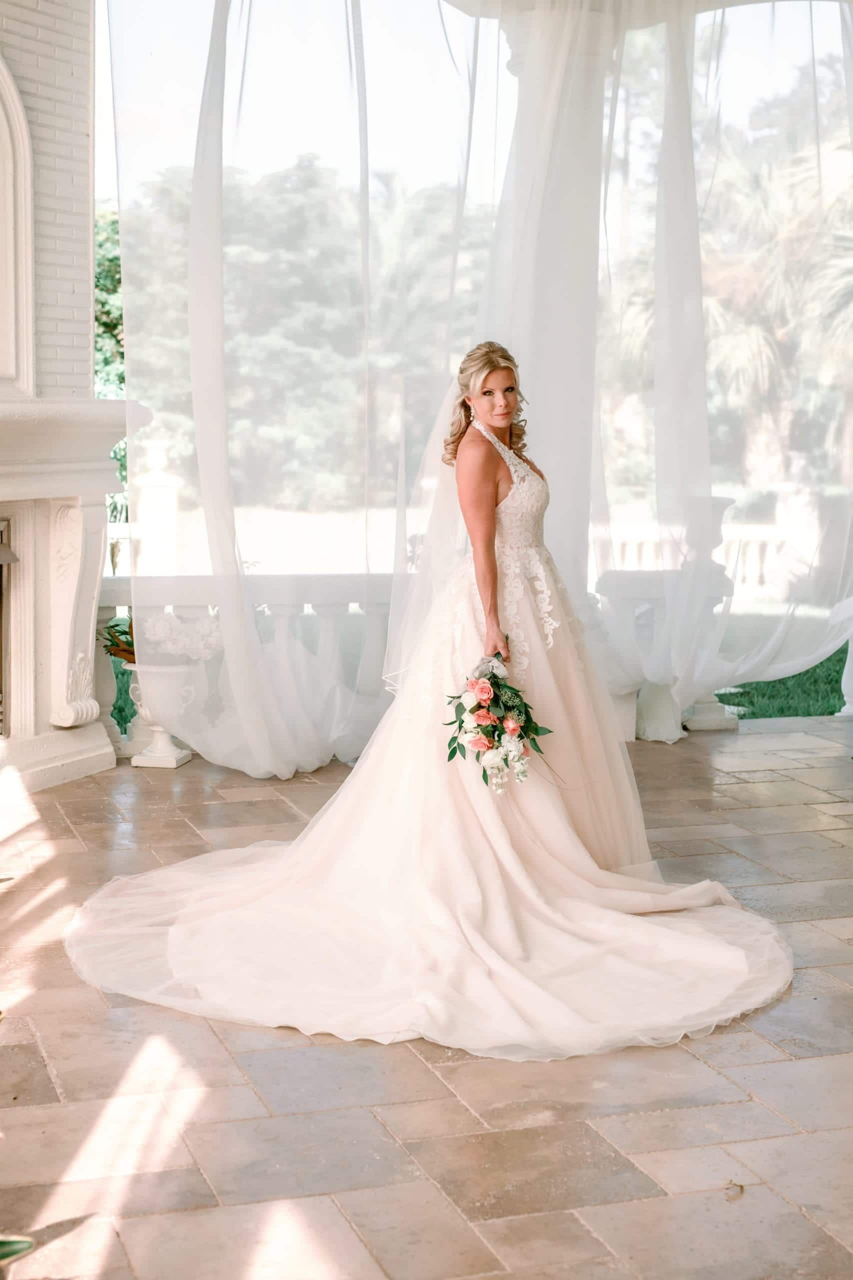 Bride in white gown, holding bouquet, smiles in a bright, airy pavilion setting.