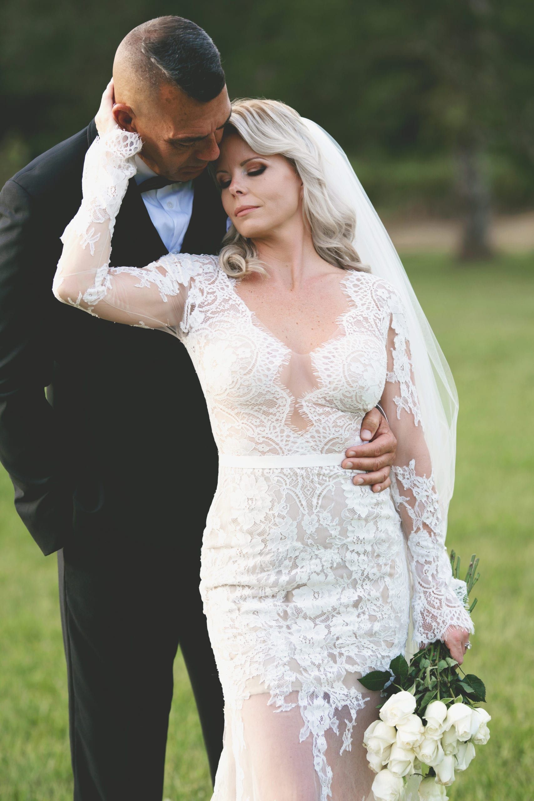 Bride and groom embrace outdoors. Bride in lace gown, groom in suit. Green background, tender moment.