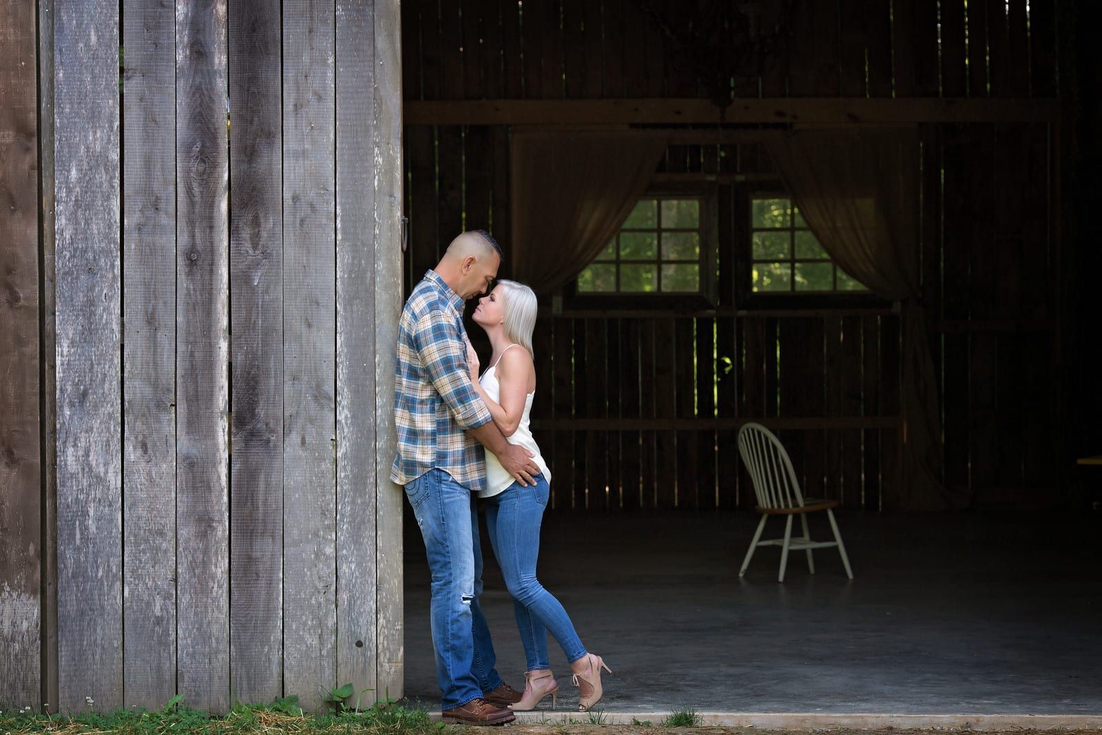Couple embracing in a barn doorway. Man in plaid shirt, woman in white top and jeans. Rustic setting, light and shadow.