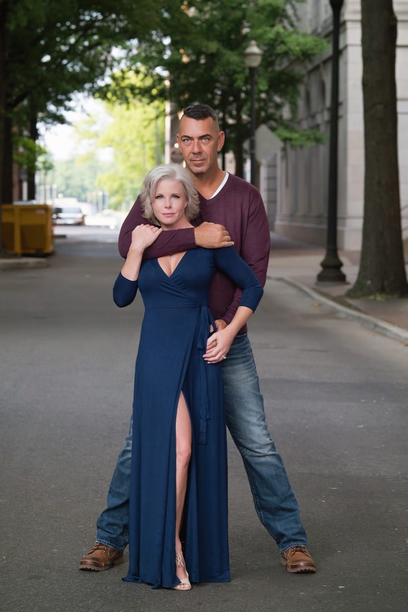 Man embraces woman from behind on a street; she wears a blue dress, he a burgundy sweater.