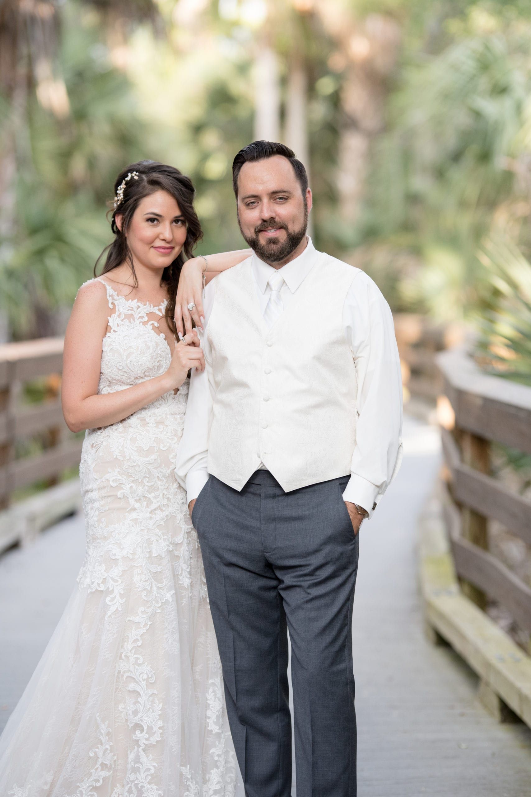 Bride and groom pose on a wooden boardwalk, smiling. The bride wears a lace wedding dress, and the groom wears a white vest.