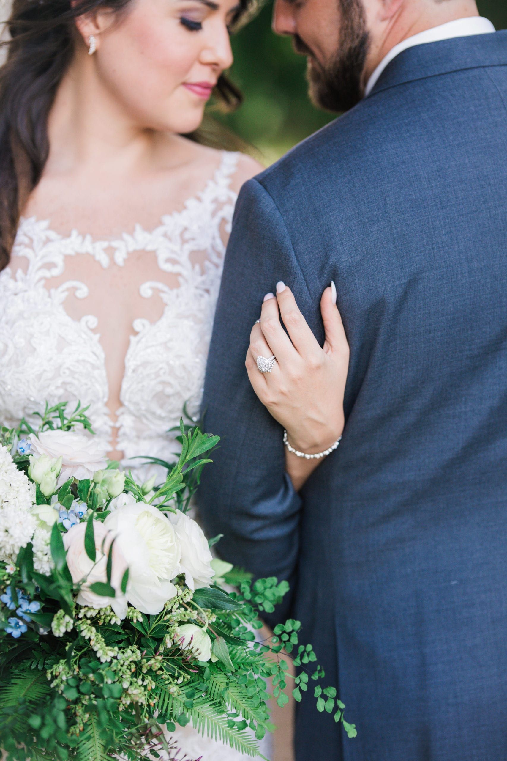 Bride and groom embrace, bride's hand on groom's suit, bouquet of white flowers.