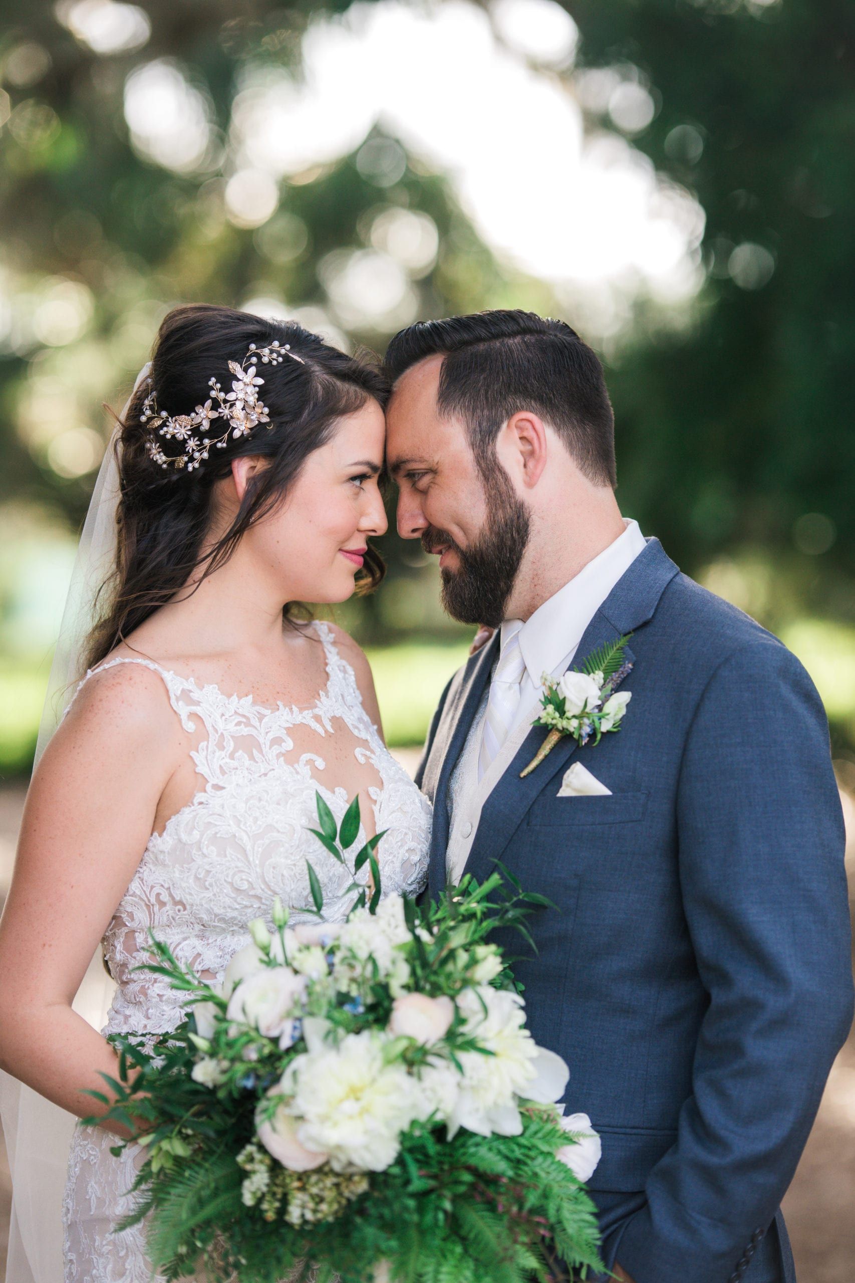 Wedding couple with foreheads touching, smiling. Bride in lace dress, holding bouquet; groom in blue suit, outdoors.