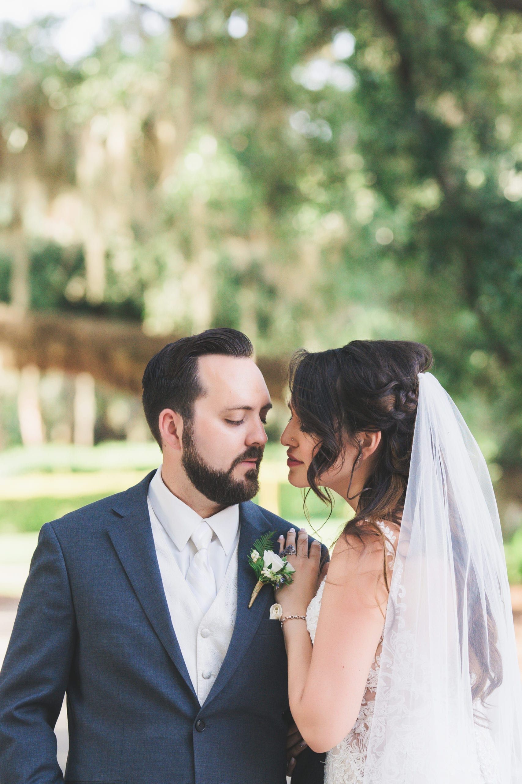 Newlyweds embrace, outdoors. Groom in blue suit, bride in wedding dress and veil, facing each other, garden backdrop.