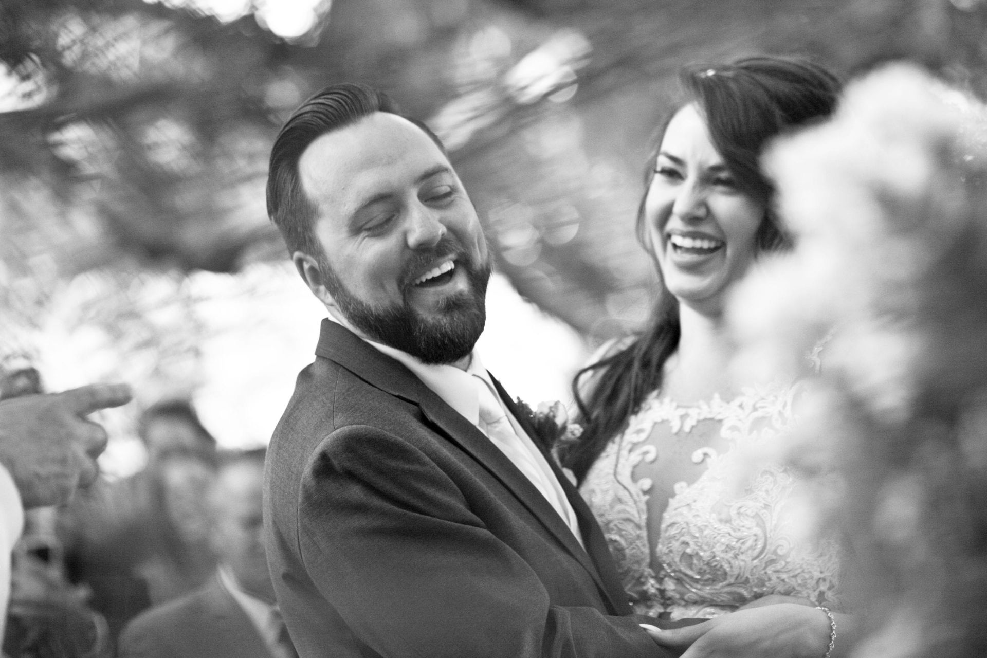 Newlyweds laughing, embracing outdoors, bride in lace gown, groom in suit.