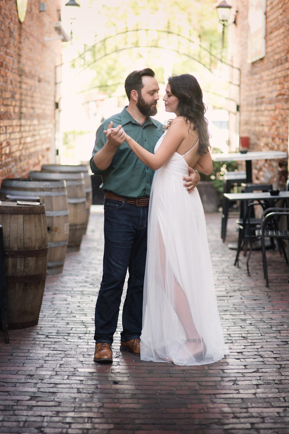Couple dancing in cobblestone alley, woman in white dress, man in green shirt, barrels and tables in background.
