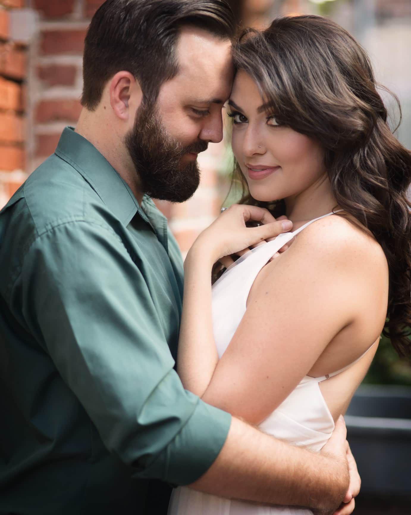 Man in green shirt embraces woman in white dress. They are close, outdoors, with a brick wall in the background.