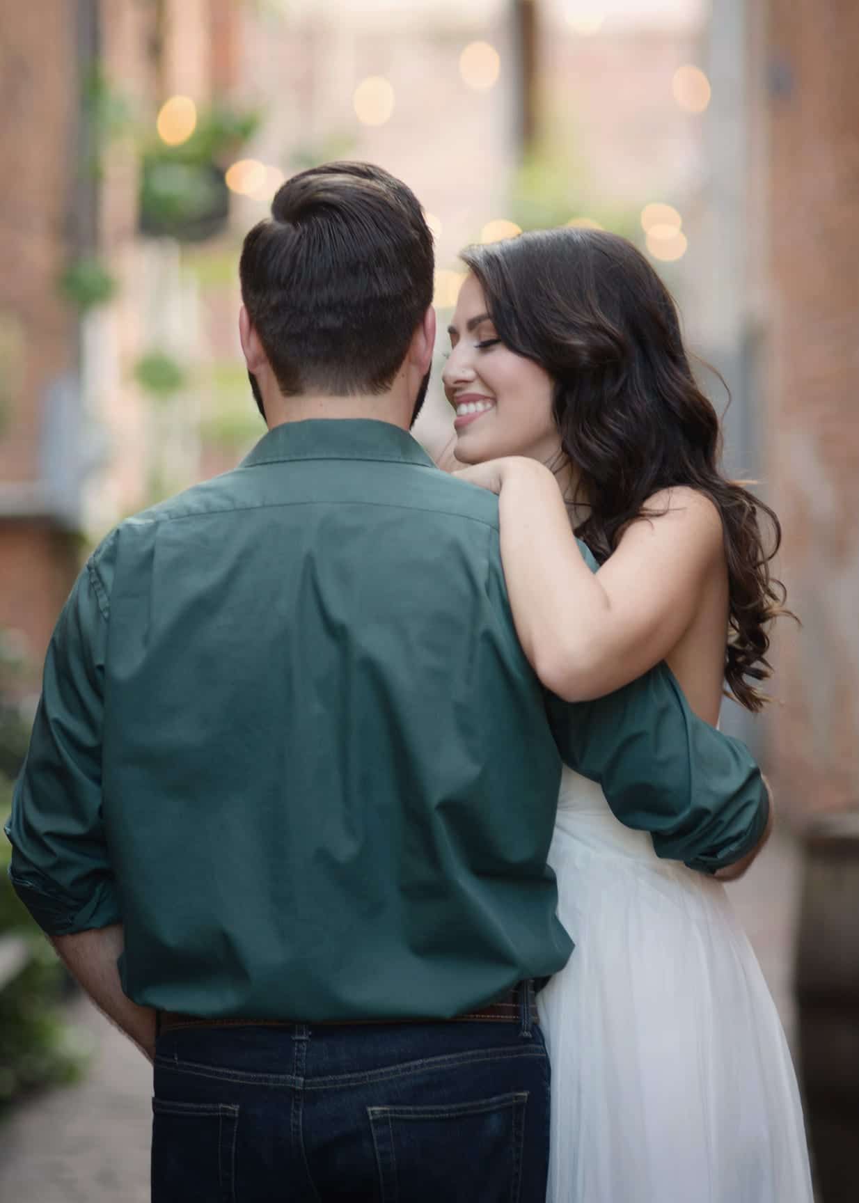 Woman smiles at man's back, arm around his shoulder, in narrow alley with string lights.