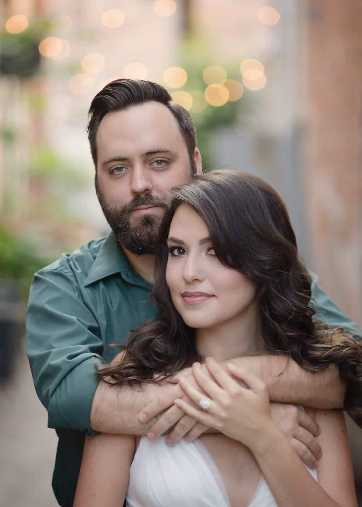 Couple embraces in an alleyway. Man in green shirt hugs woman in white dress. Soft bokeh background.
