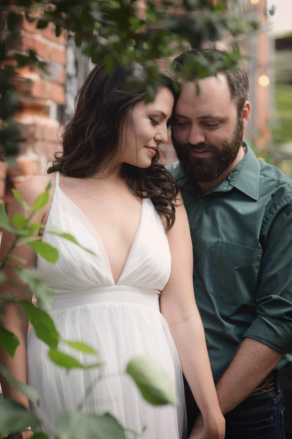 Woman in white dress and man in green shirt embracing, smiling. Outdoor setting.