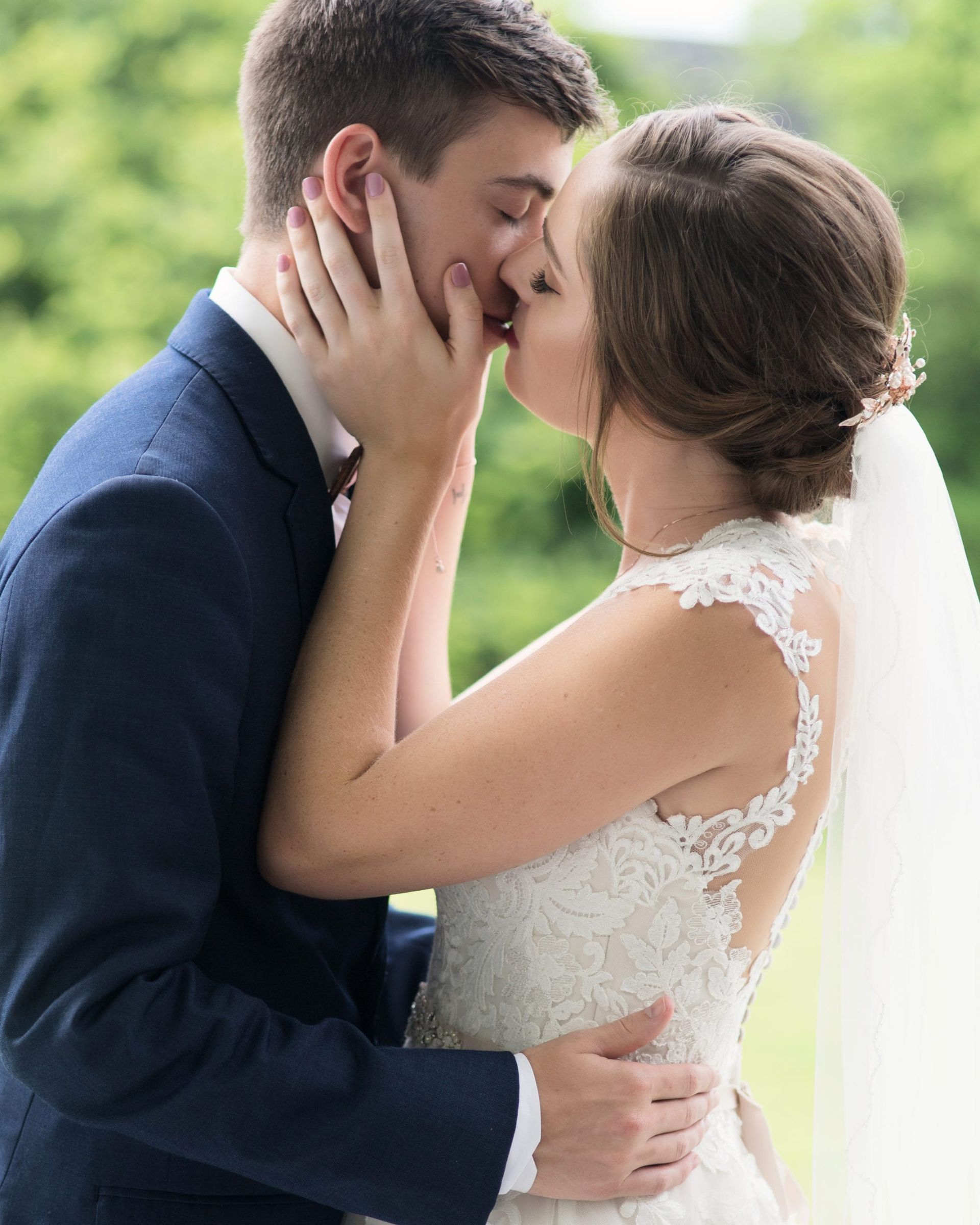 Bride and groom kissing, hands on faces; outdoor wedding, green background.