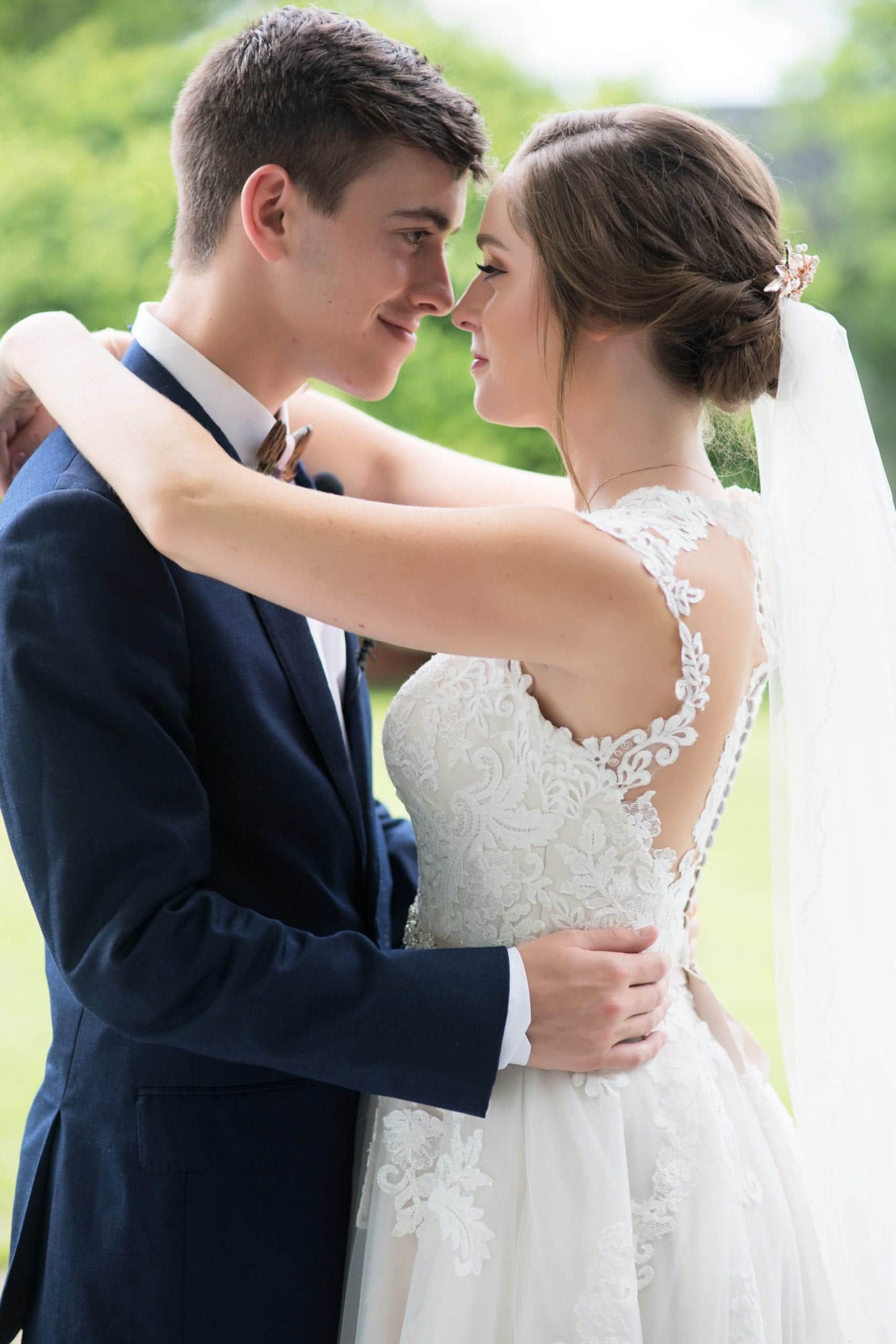 Groom in navy suit embracing bride in white lace dress; both smiling, outdoors.