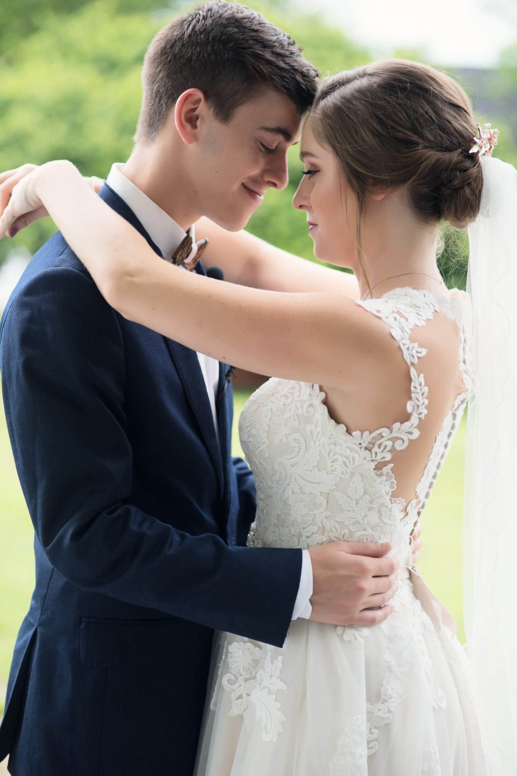 Bride and groom embracing; he's in a suit, she's in a white dress. Smiling, close, soft focus, outdoor setting.