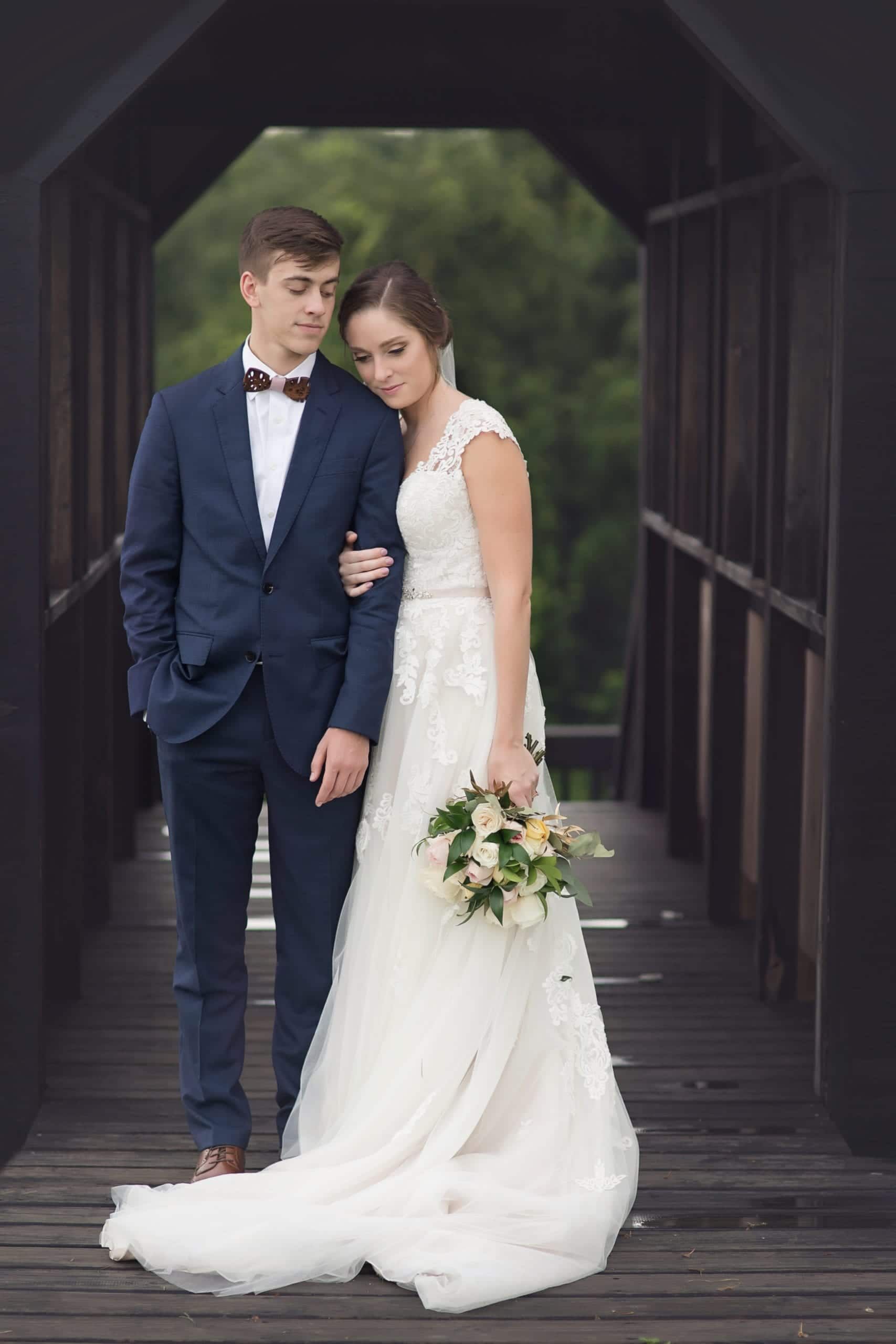 Groom in blue suit and bride in white gown on a wooden bridge, embracing. Bride has a bouquet, both look down.
