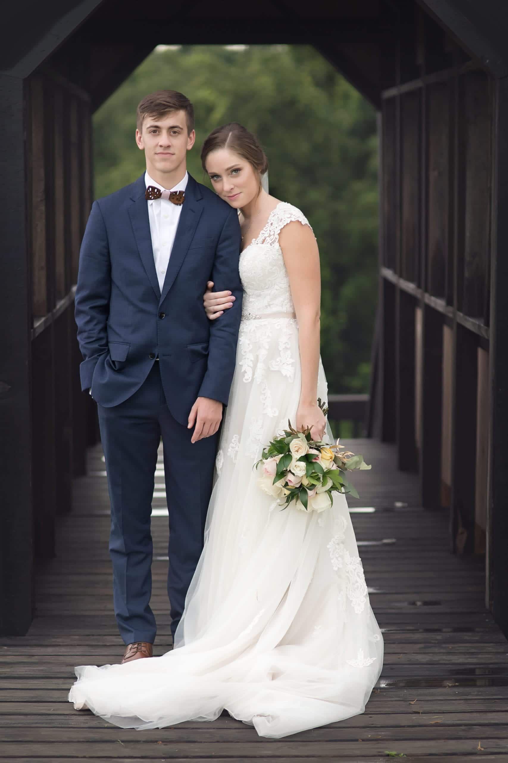 Young couple on a wooden bridge. The woman wears a wedding dress, while the man is in a suit. They pose together.