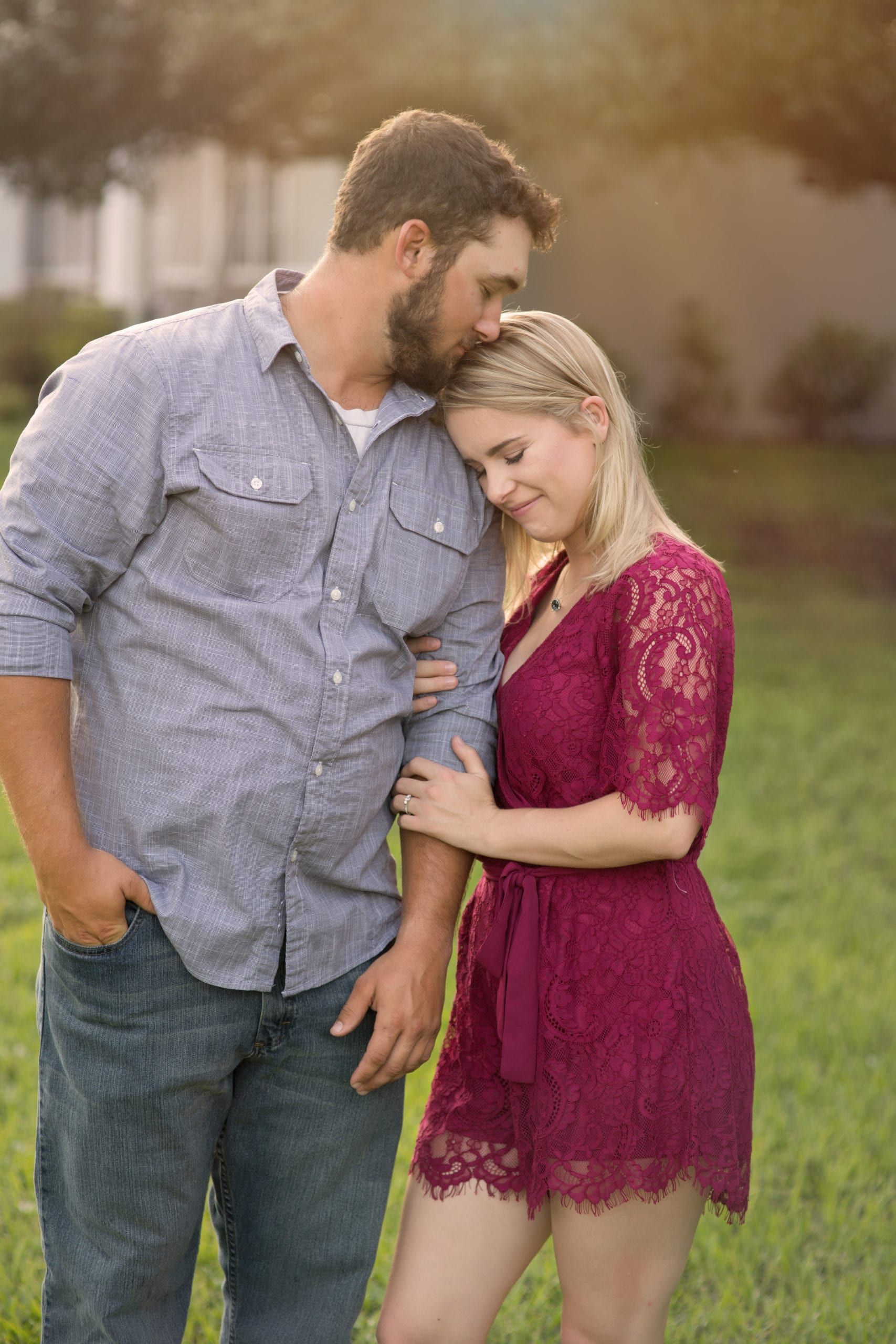 Man kissing woman's forehead; couple in a park, man in a blue shirt and jeans, woman in a red dress.
