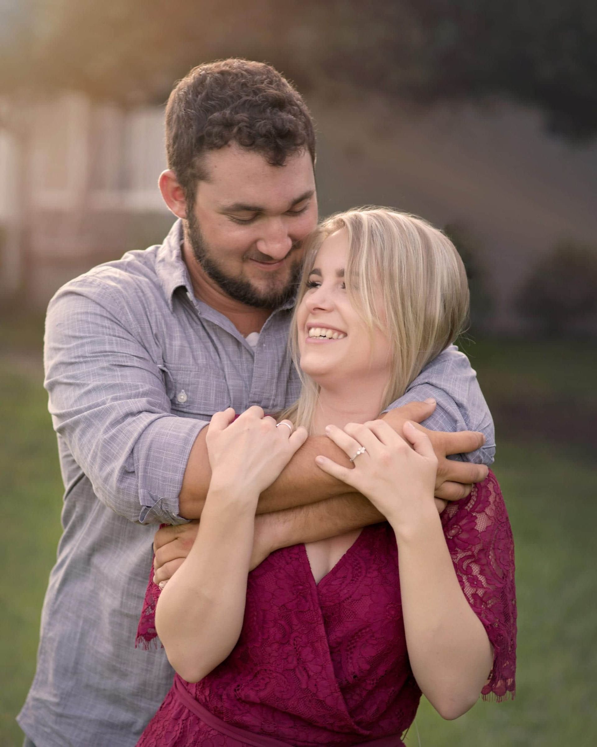 Man hugging woman, both smiling. Man wears blue shirt, woman wears red dress; outdoors.