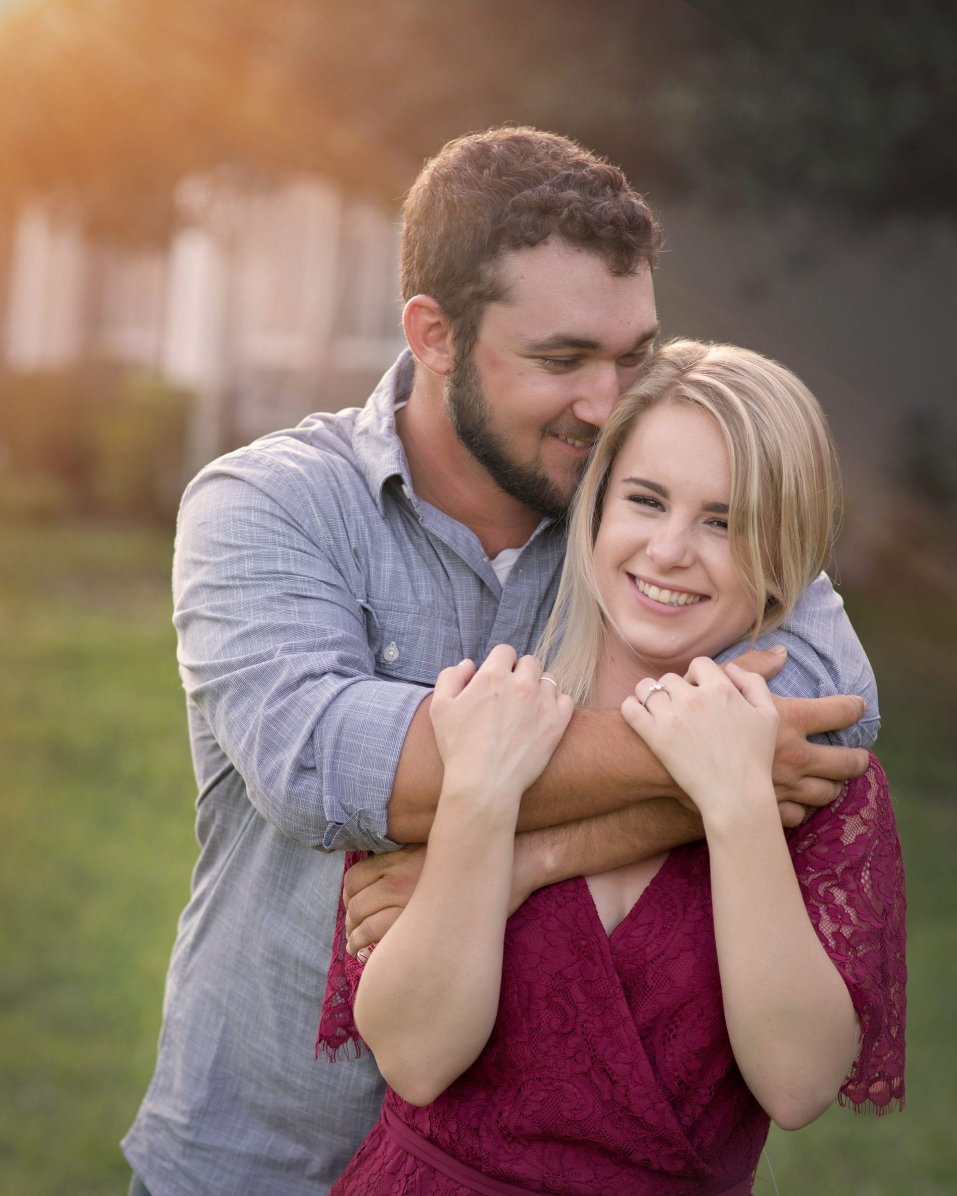 Man embraces woman, both smiling, outdoors. He's in a blue shirt; she's in a red dress.