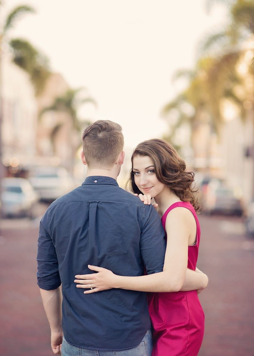 Woman in red dress hugs man in blue shirt on a brick road, looking over shoulder with a smile.
