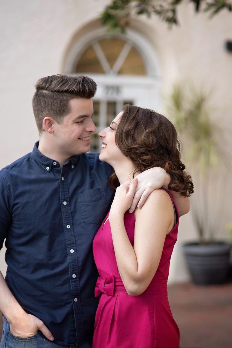 Man in blue shirt embraces woman in pink dress, smiling, in front of a building.