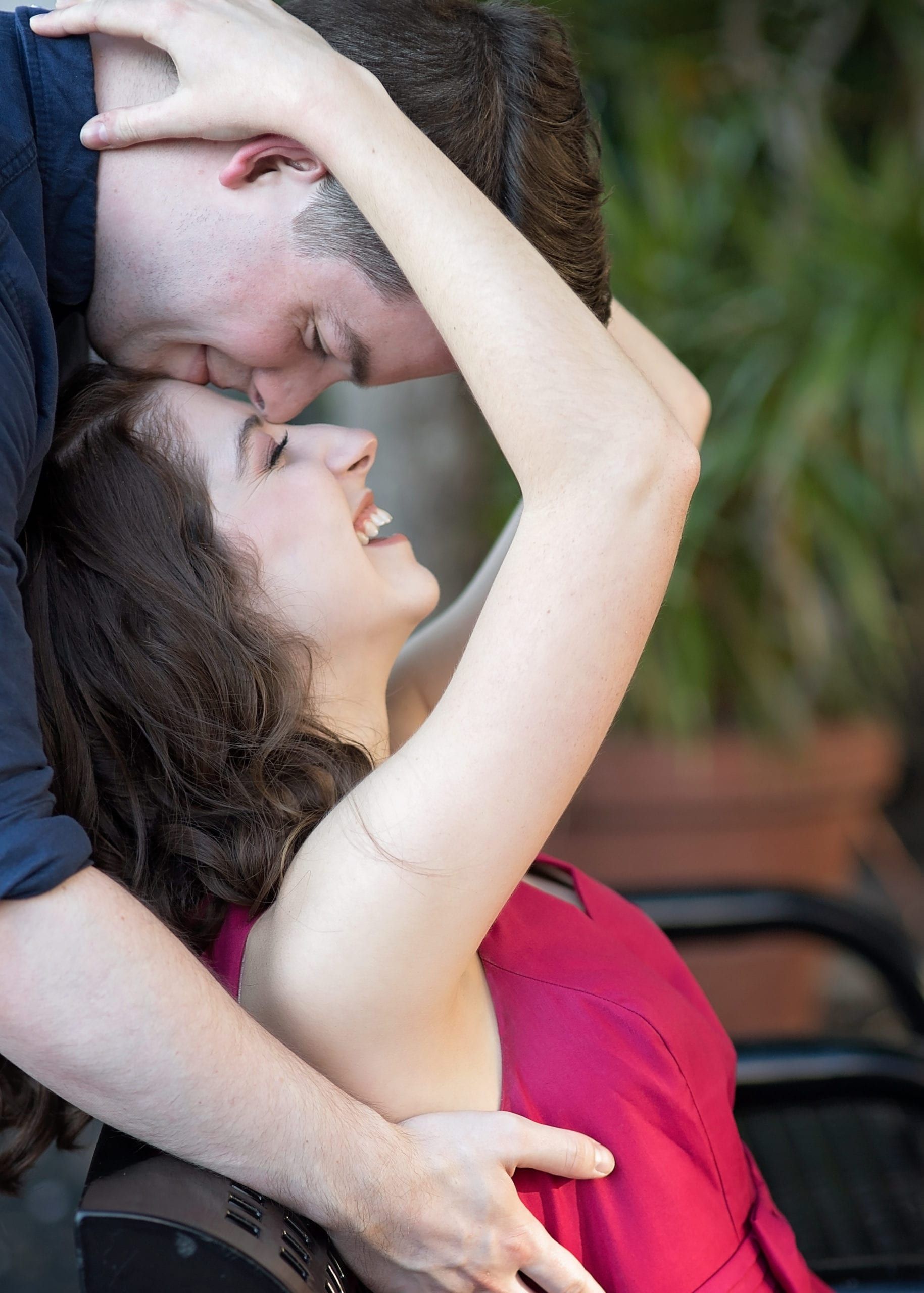 Man embracing woman, kissing her forehead. She laughs, wearing a pink dress, outdoors.