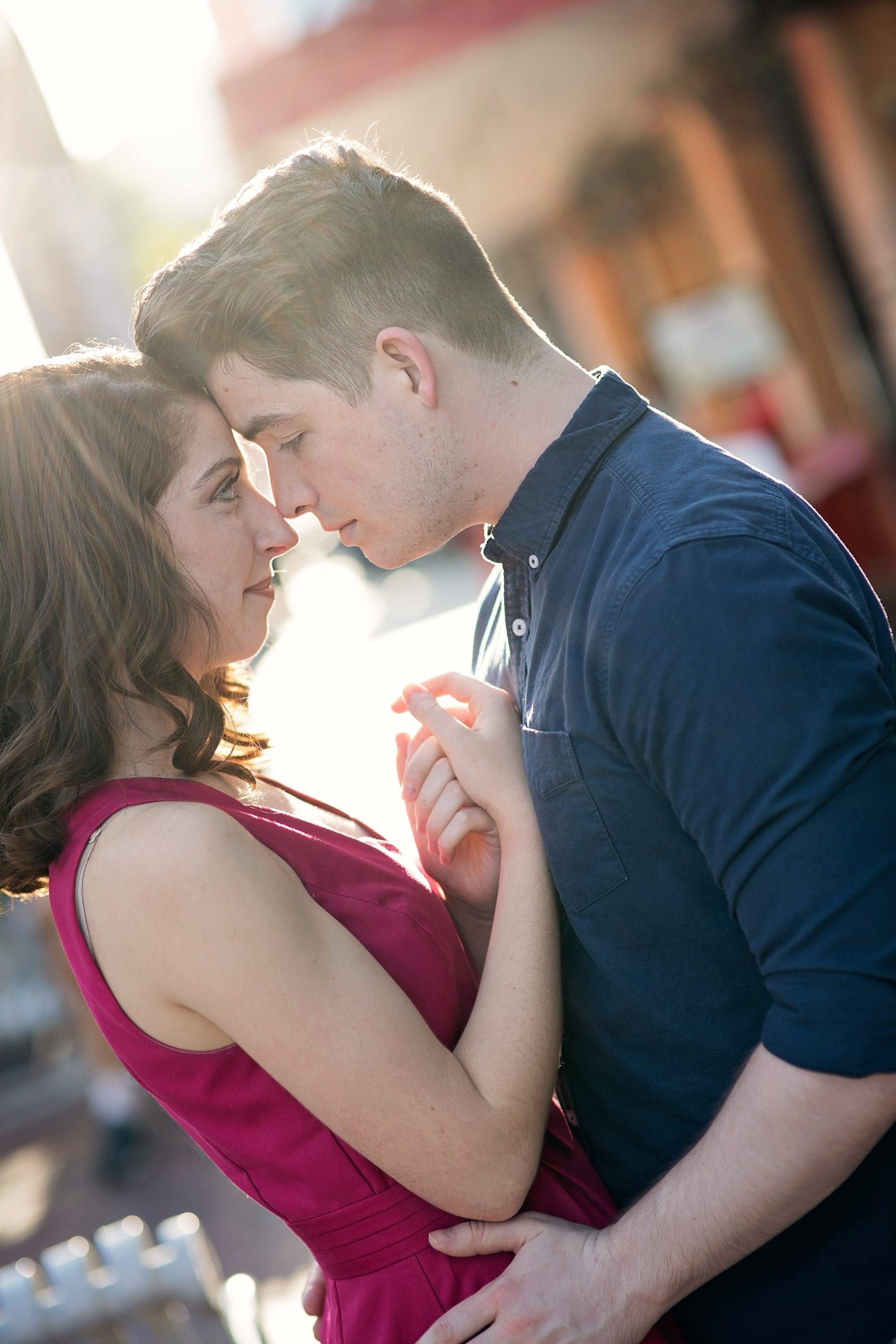 Couple embracing, holding hands, looking at each other. Woman in red dress, man in blue shirt, outdoors.
