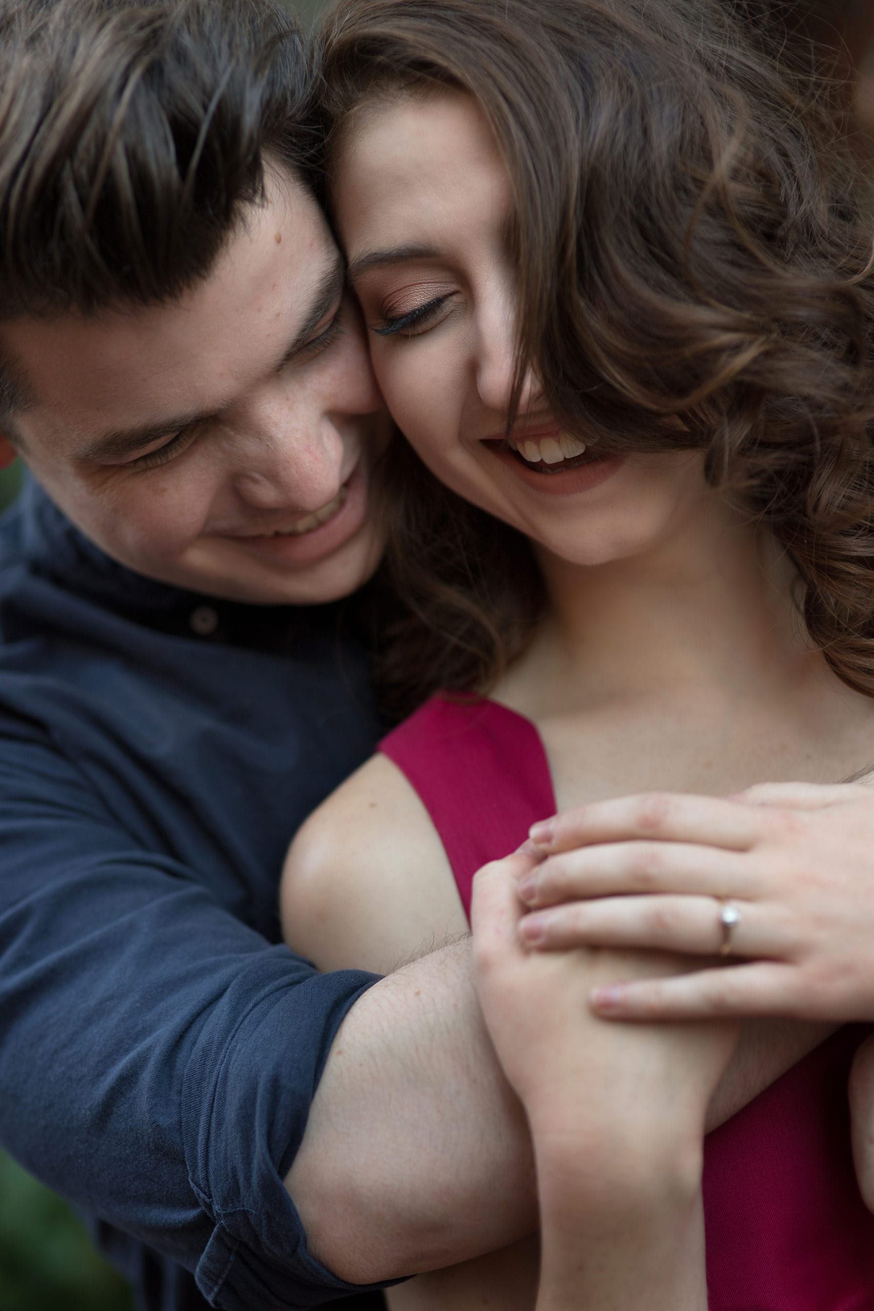 Man hugging woman, both smiling, outdoors. Woman wearing red dress, man in blue shirt.