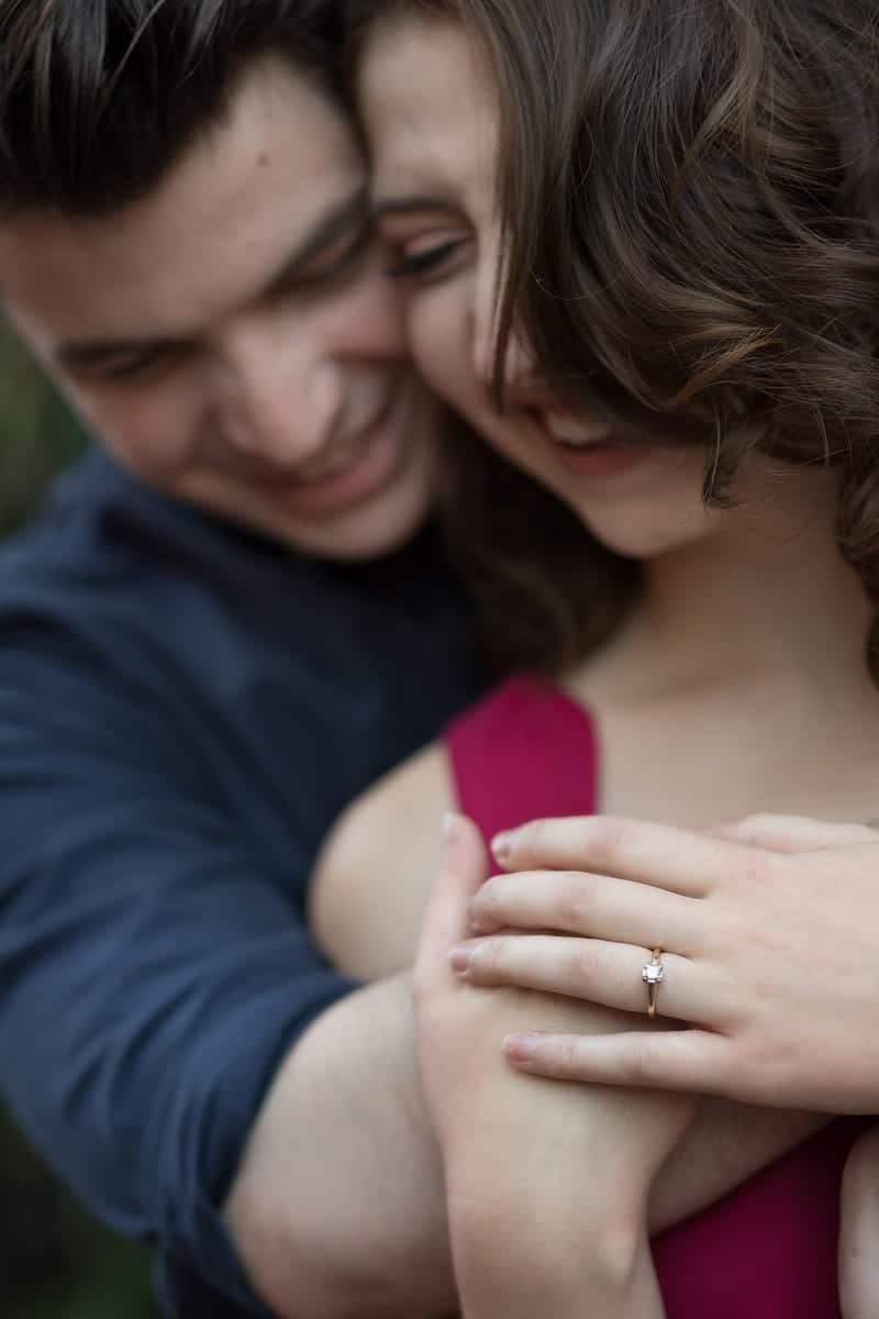 Man embracing woman; both smiling. She wears a ring, red dress. Outdoors.
