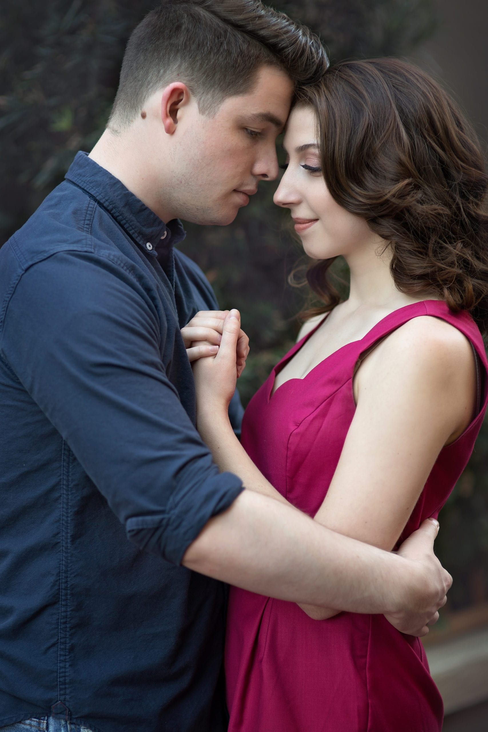 Couple embraces, foreheads touching, holding hands. Man in blue shirt, woman in pink dress, outside.