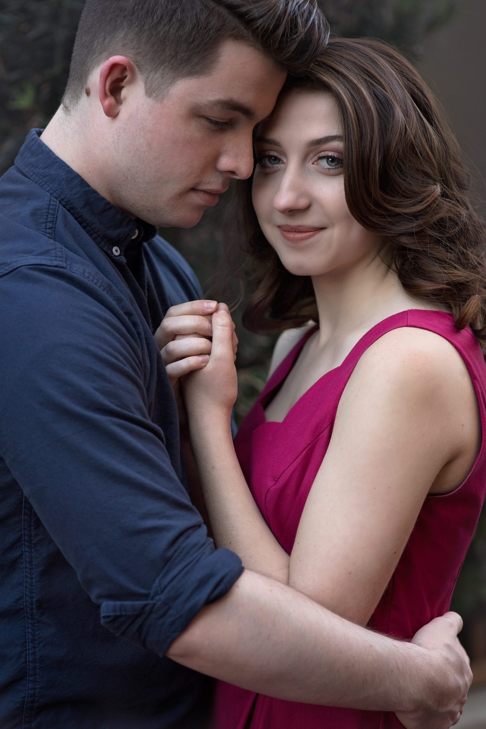 Couple embraces; woman in pink dress smiles, man in blue shirt looks down. Outdoors.