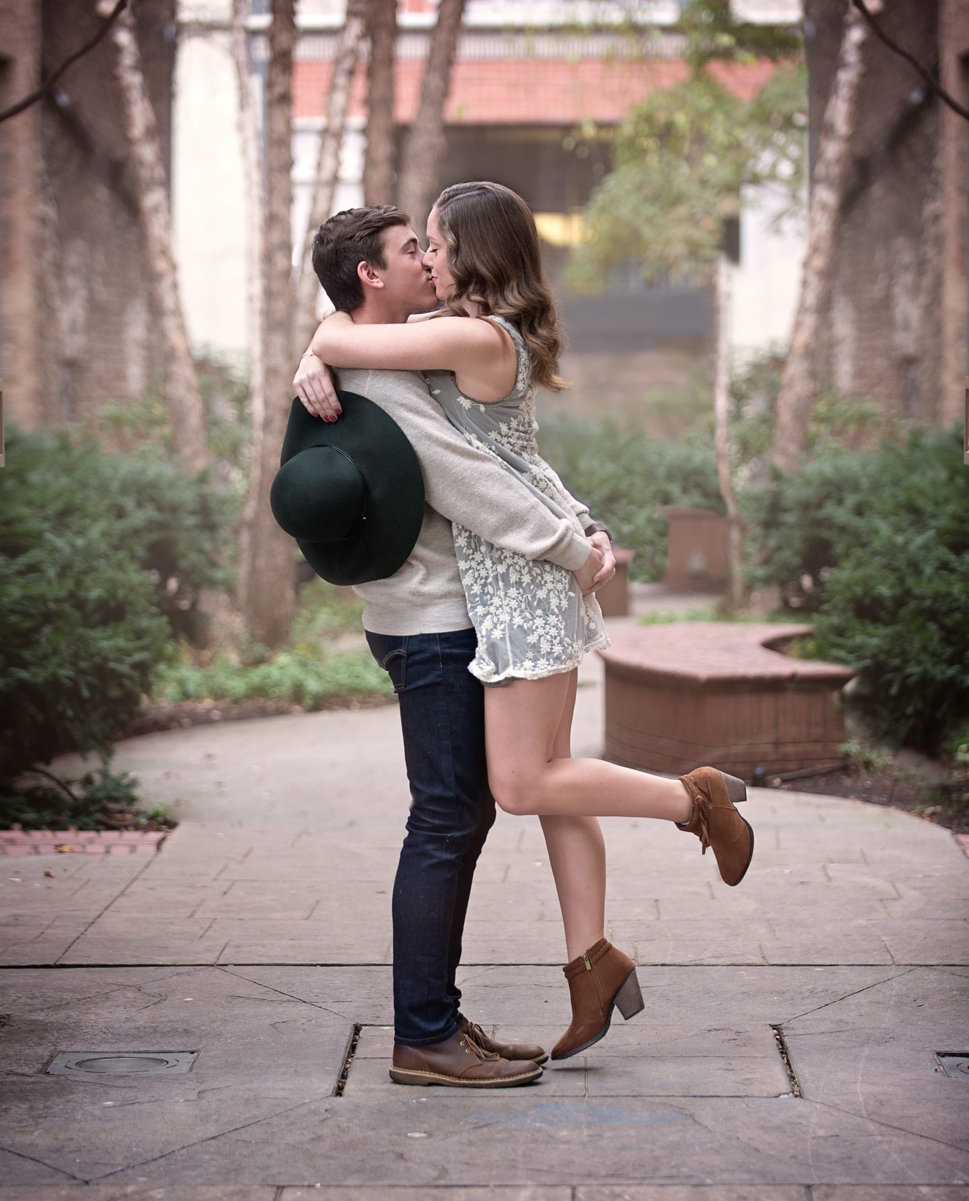 Man lifts woman, kissing, in outdoor setting. She wears a floral dress and boots; he wears jeans and holds a hat.