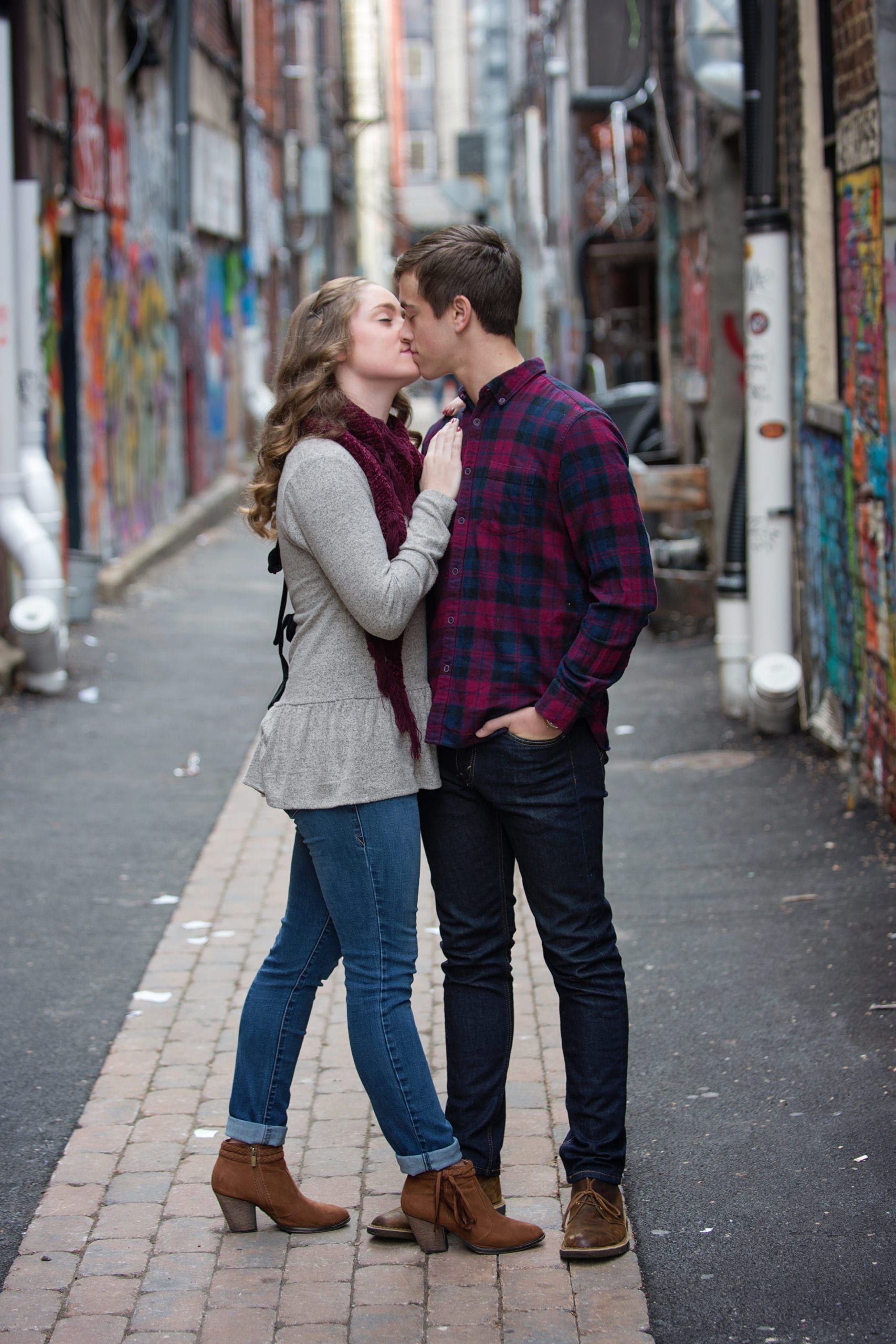 Couple kissing in a brick-paved alley with graffiti art. Woman in scarf, jeans, and boots, man in plaid shirt.