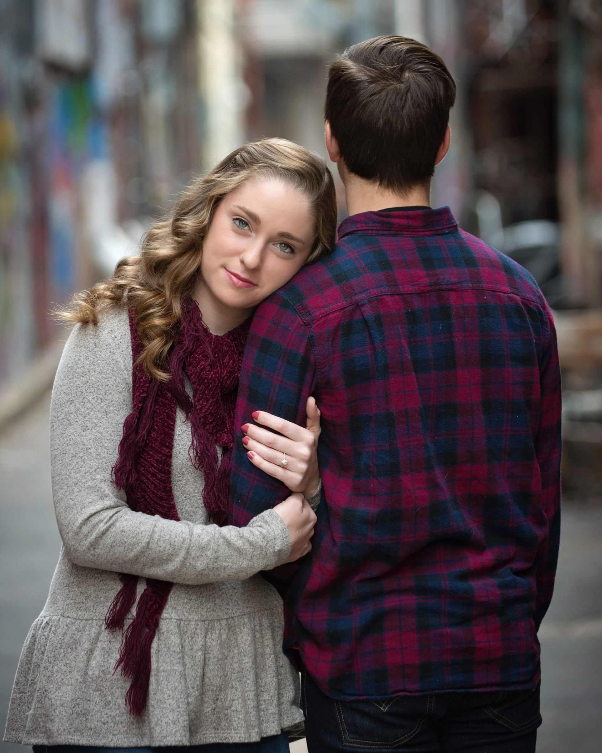 Woman leans head on man's back in an alley. She wears a gray sweater, scarf. He has on a plaid shirt.