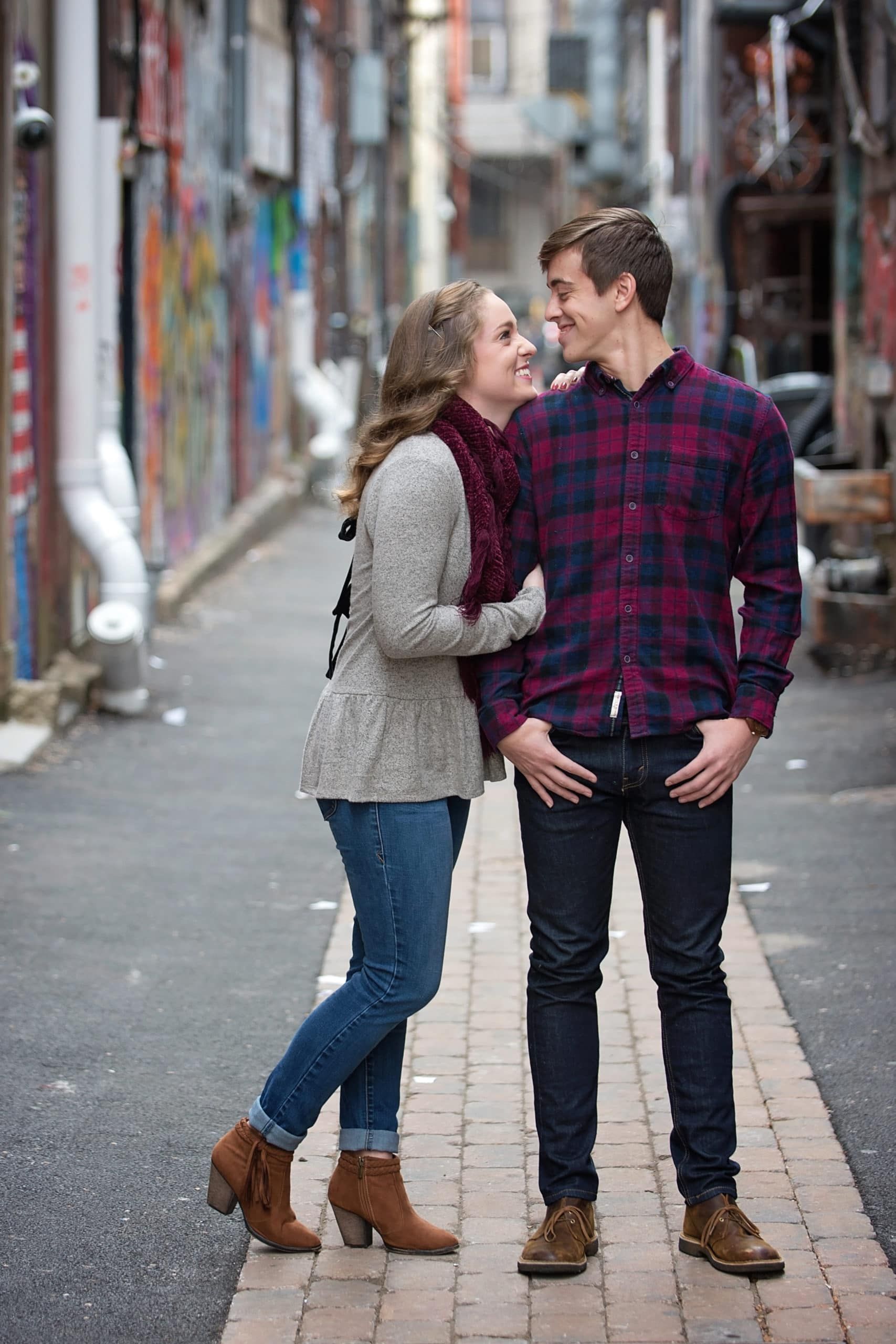 Couple smiling at each other in alleyway, the woman has her arm on the man's shoulder. Graffiti and brick.