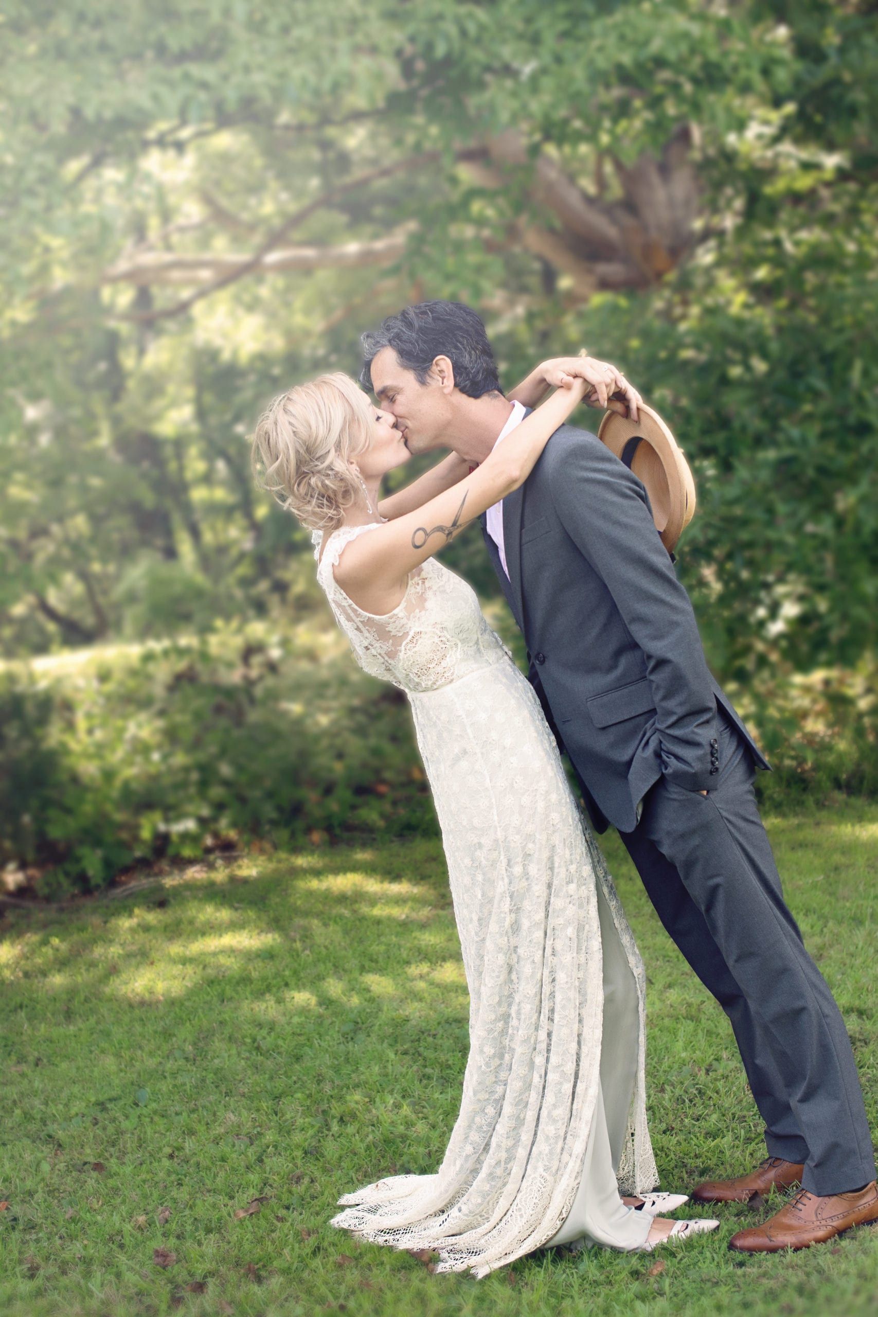 Bride and groom kissing in outdoor setting; she leans back, he leans forward, embracing. Green grass, trees.