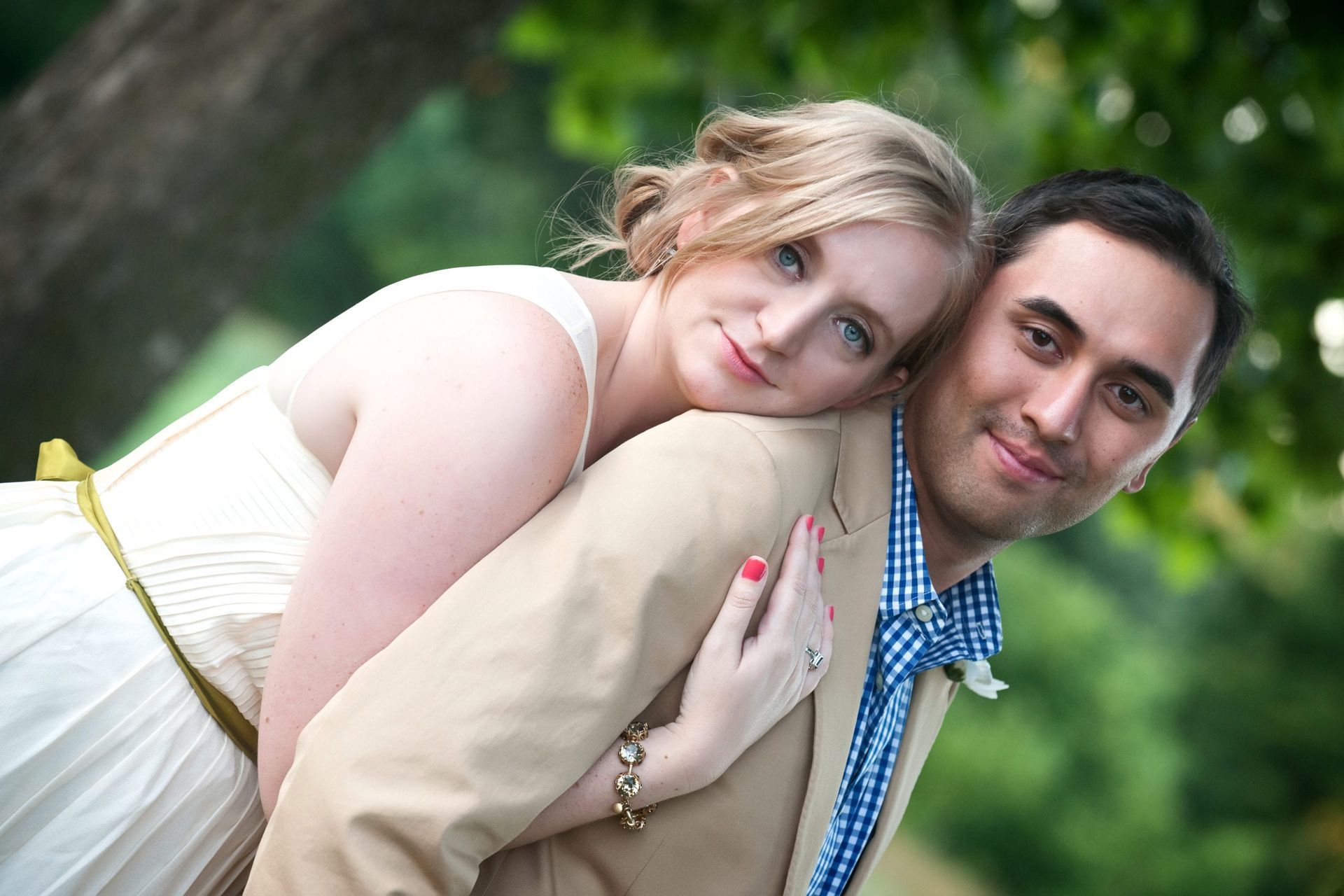 Bride leans on groom's back; both smiling in an outdoor setting. She wears a white dress, he wears a tan jacket and blue shirt.