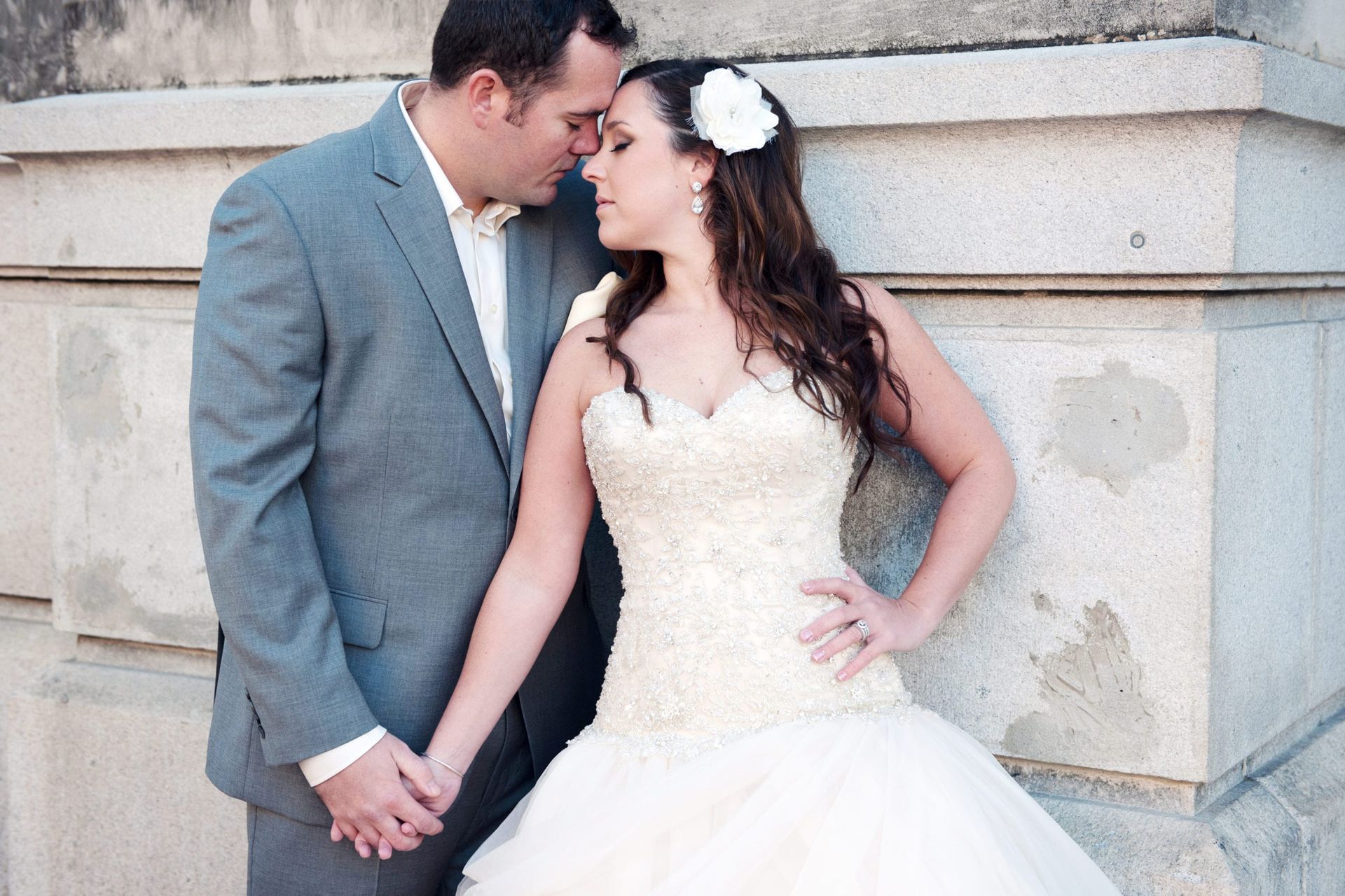 Wedding couple, man in gray suit, woman in white gown, holding hands, leaning against a stone wall.