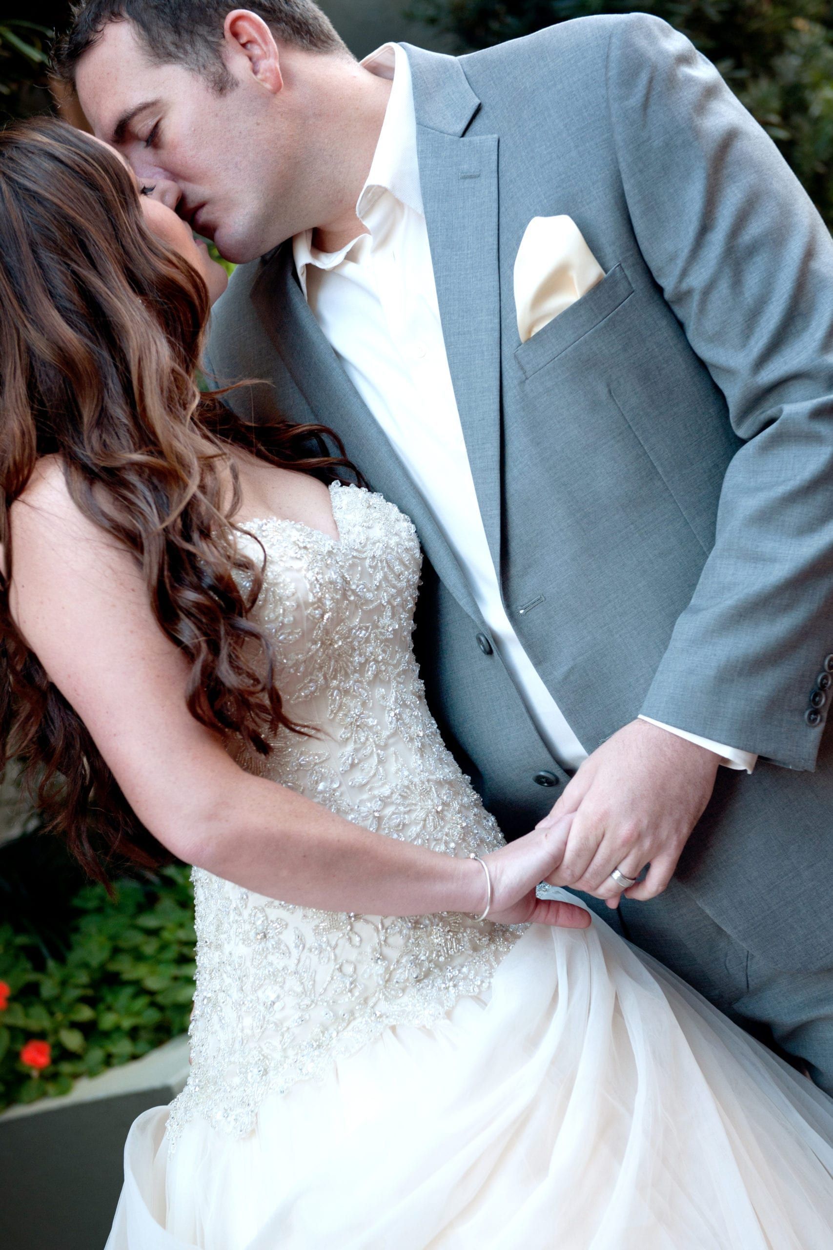 Bride and groom kissing; woman in white beaded gown, man in gray suit.