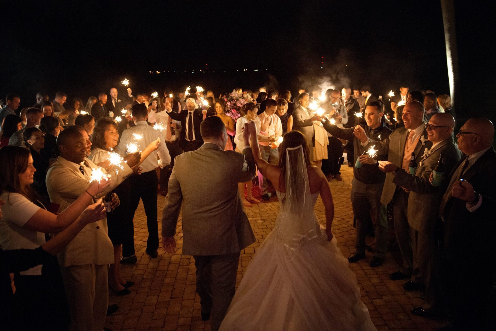 Bride and groom walk through a crowd holding sparklers at night.