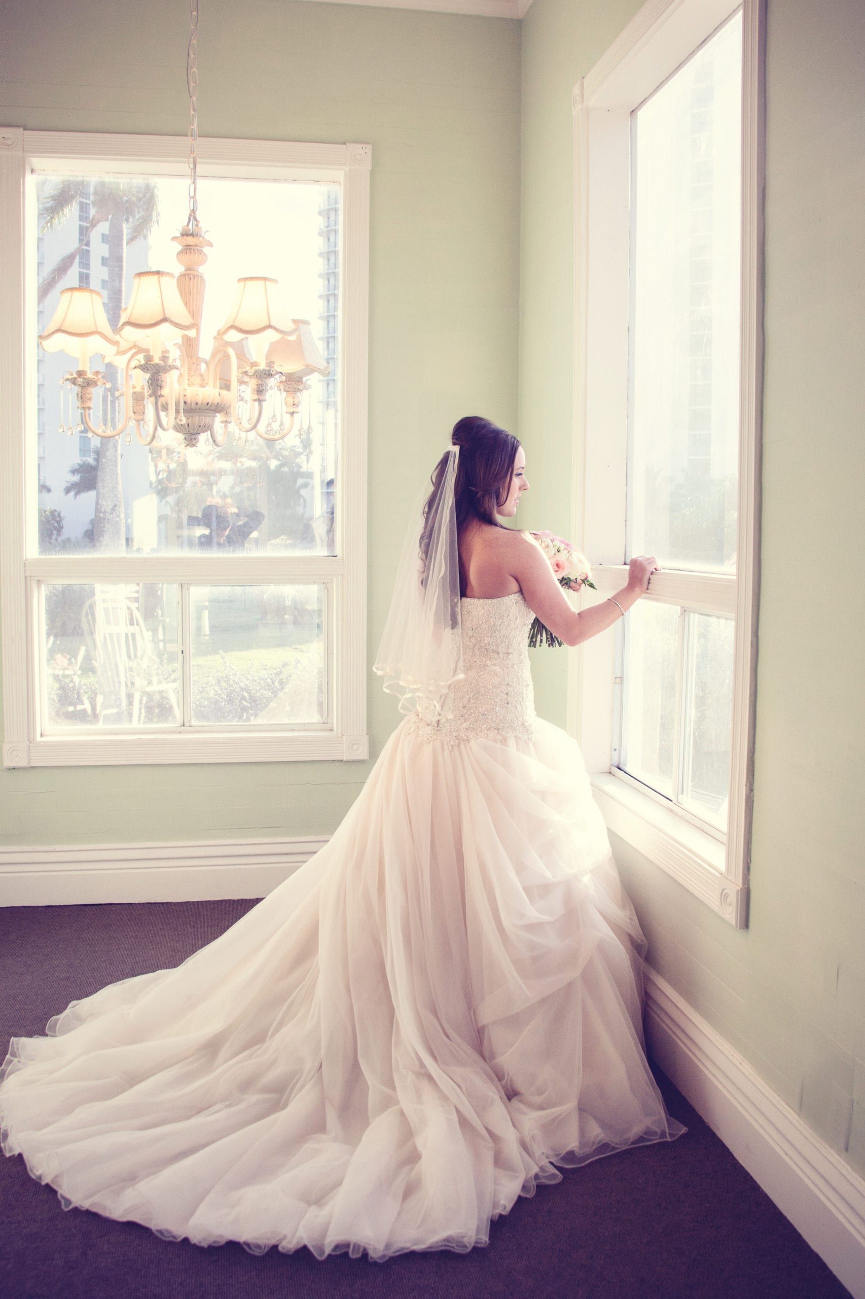 Bride in a white wedding dress looks out a window in a pastel green room.