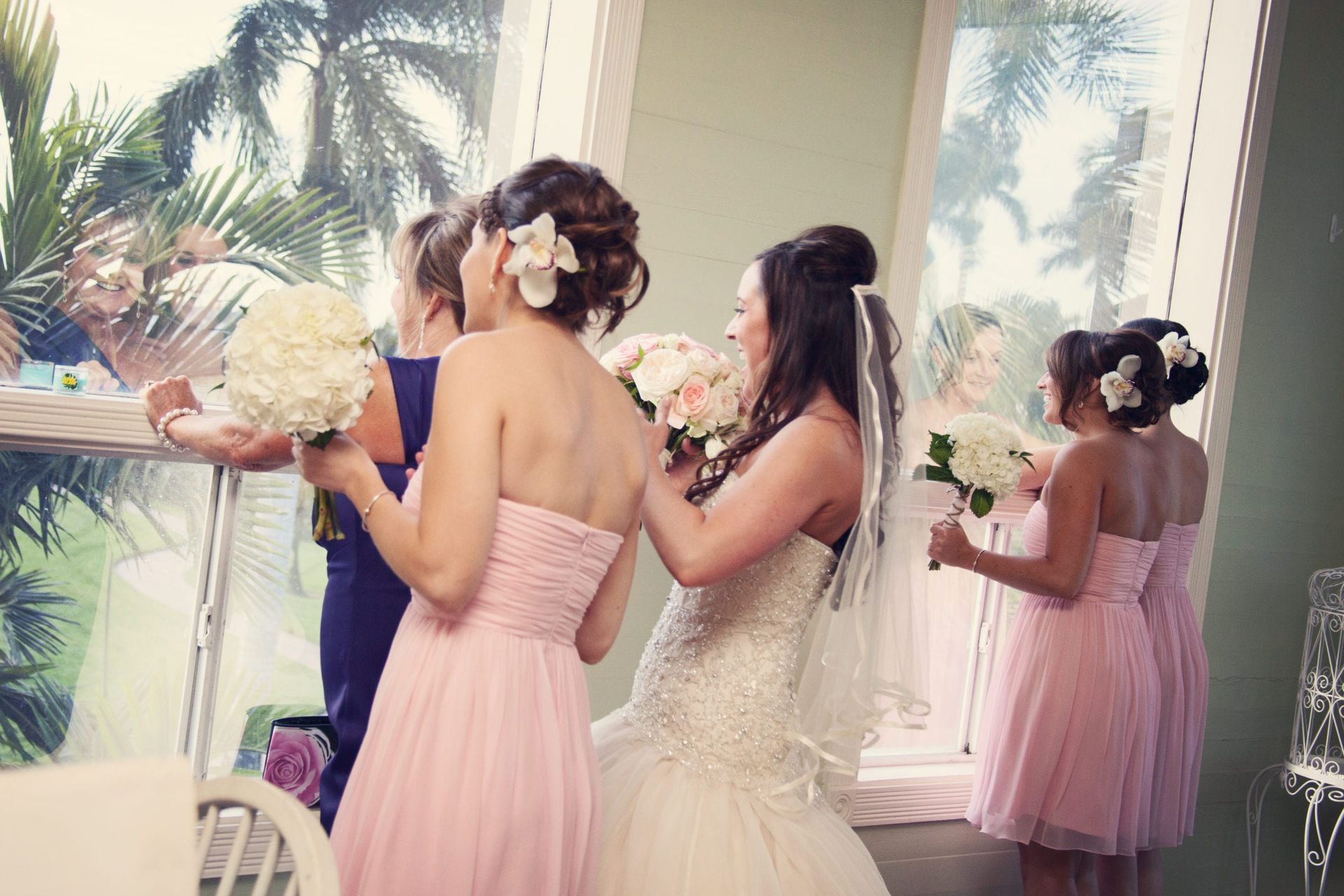Bride in white dress with bridesmaids in pink dresses, looking out a window at tropical greenery.